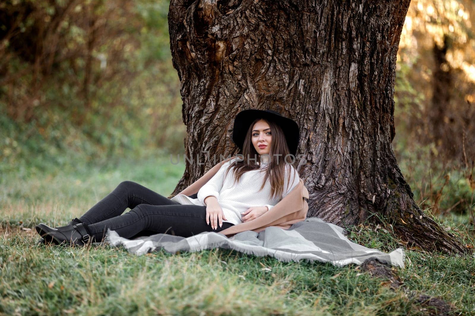 beautiful woman in a beige coat and black hat relaxing under big tree on sunny autumn day.