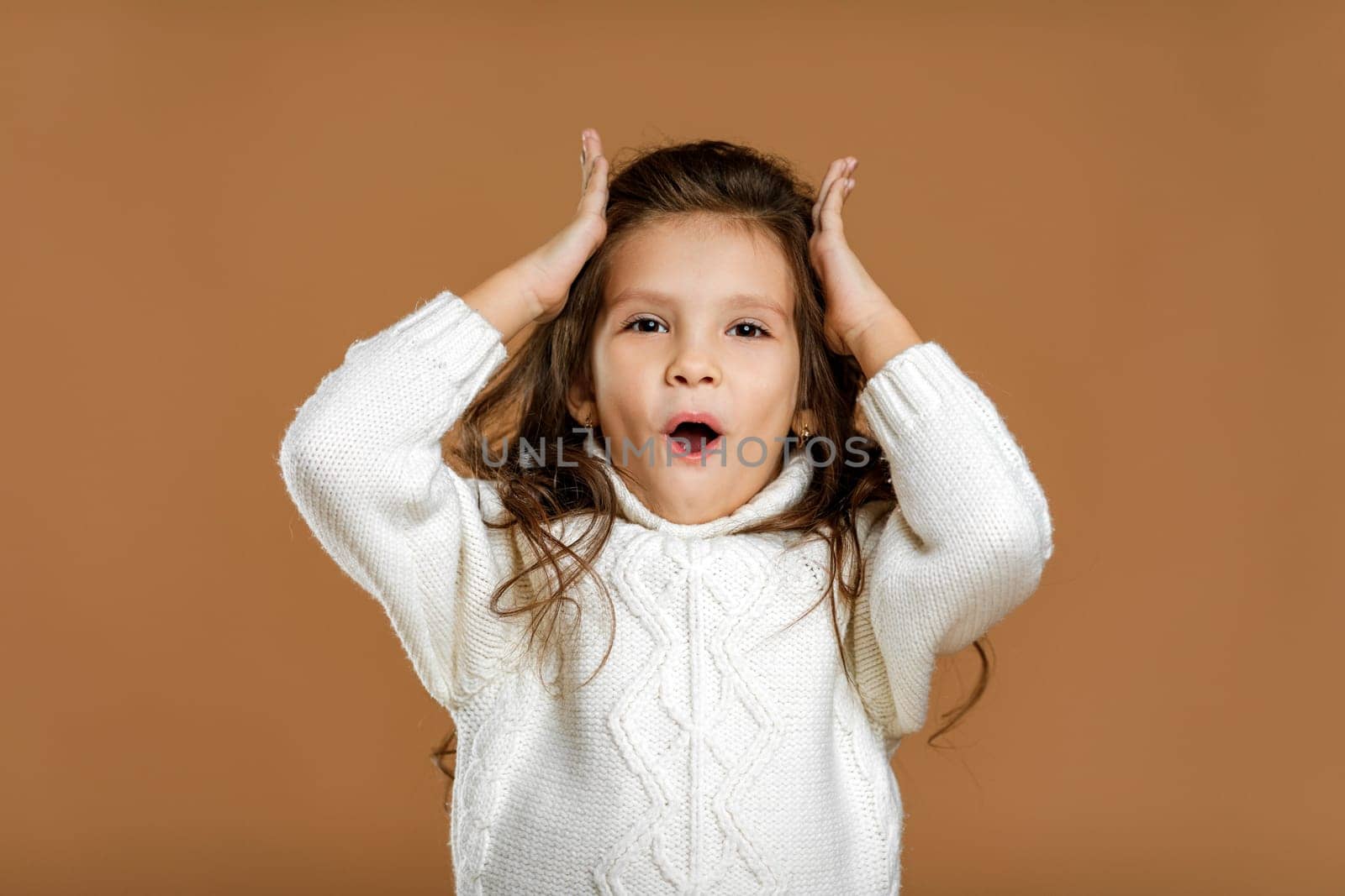 happy surprised beautiful curly little child girl in white sweater looking to camera on beige background. Human emotions and facial expression