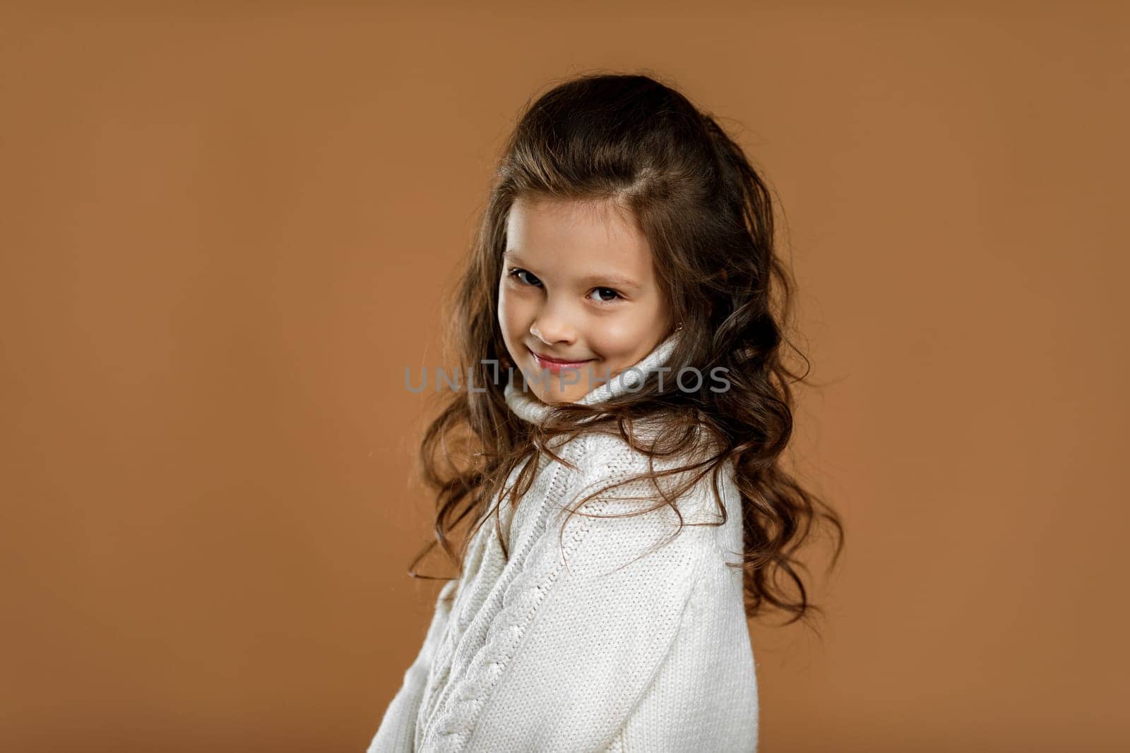 Cute shy little child girl in white sweater looking to camera on beige background. Human emotions