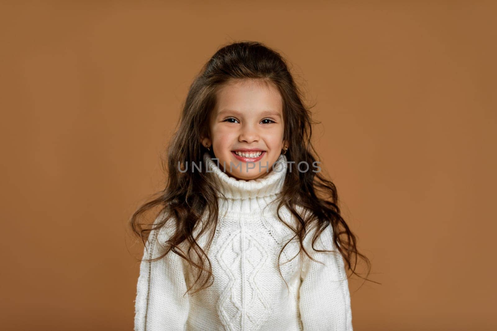 Cute smiling little child girl in white sweater looking to camera on beige background. Human emotions and facial expression