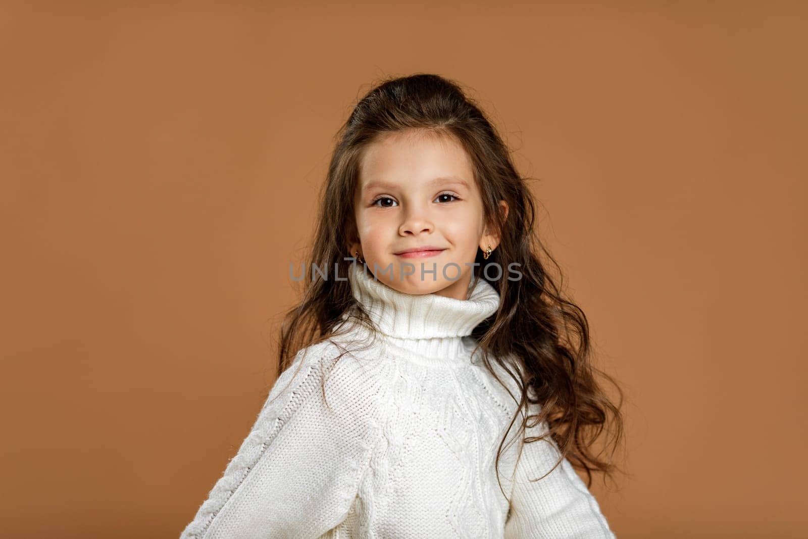 Cute smiling little child girl in white sweater looking to camera on beige background. Human emotions and facial expression