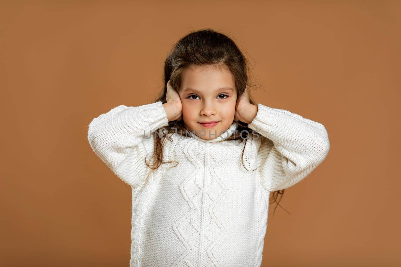 Cute curly little girl in white sweater is closing her ears with her hand on beige background.