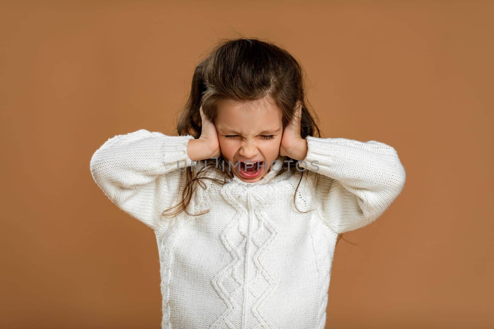 Cute curly little girl in white sweater is closing her ears with her hand and screaming on beige background.