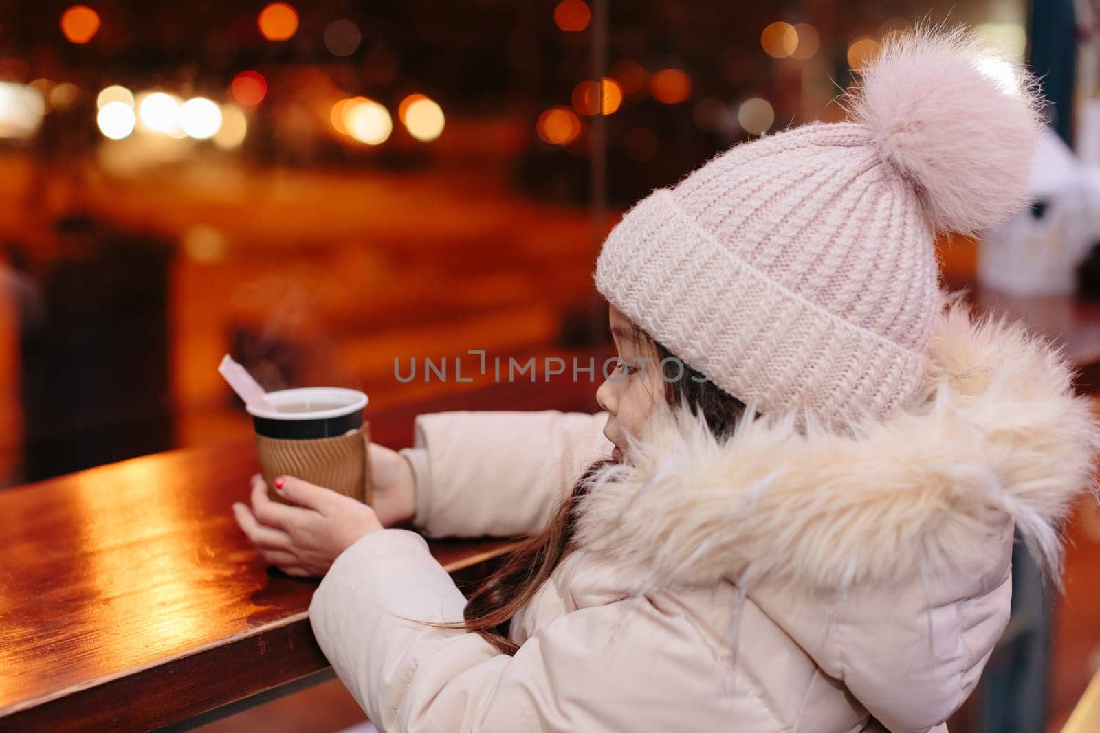 Cute happy little girl drinks hot tea in a street cafe in the evening in winter. child is cold. Christmas holidays