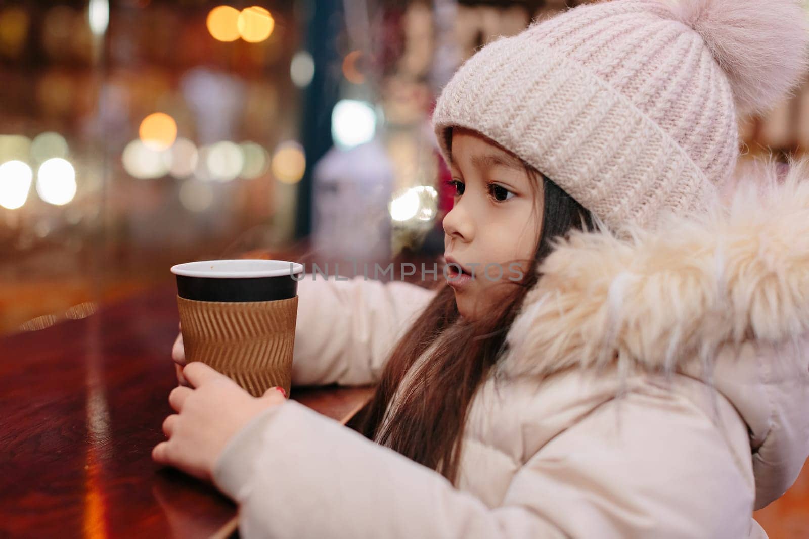 Cute happy little girl drinks hot tea in a street cafe in the evening in winter. child is cold. Christmas holidays