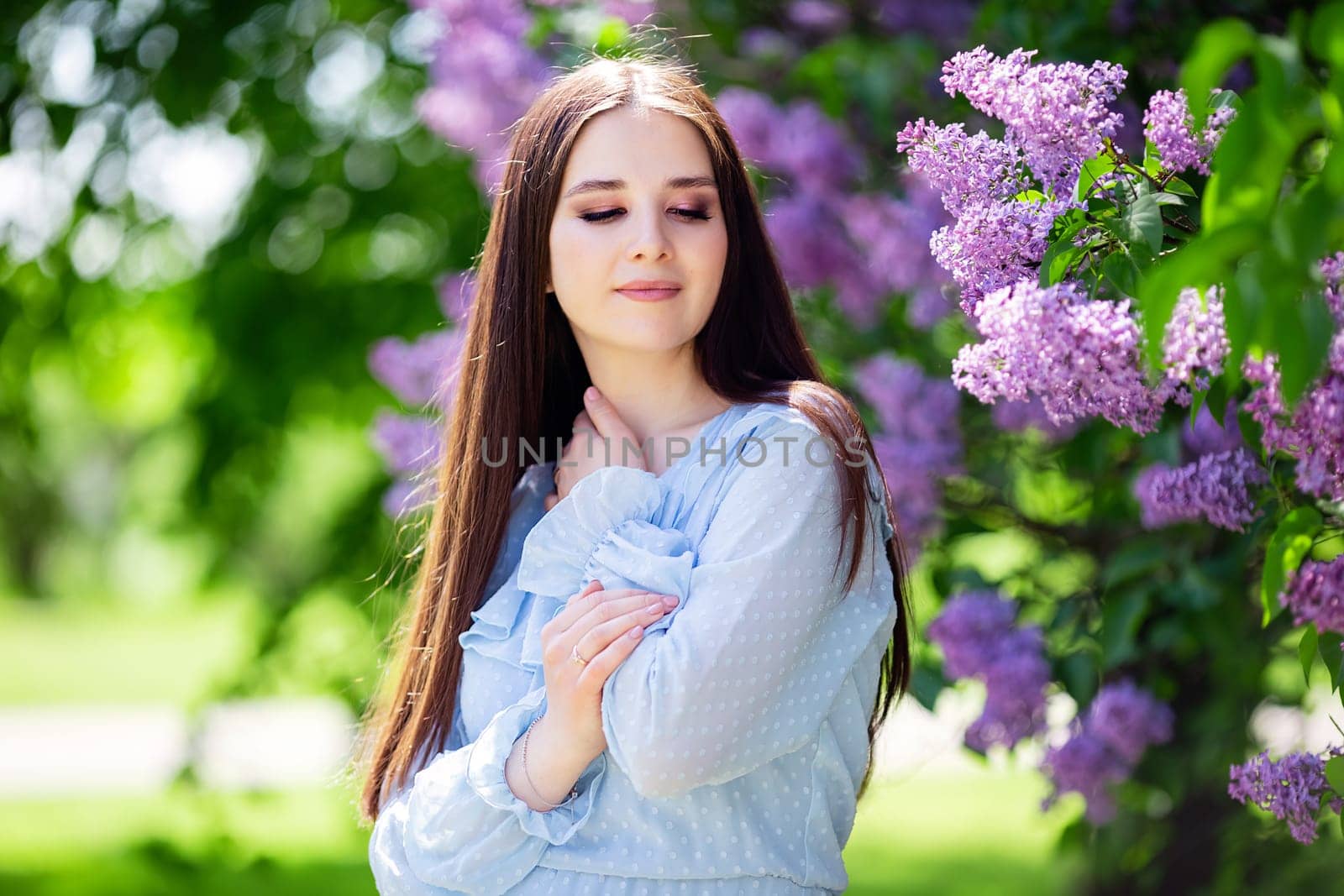 A beautiful enjoying girl in light blue dress, stands next to a flowering lilac bush in the park. Close up. Copy space
