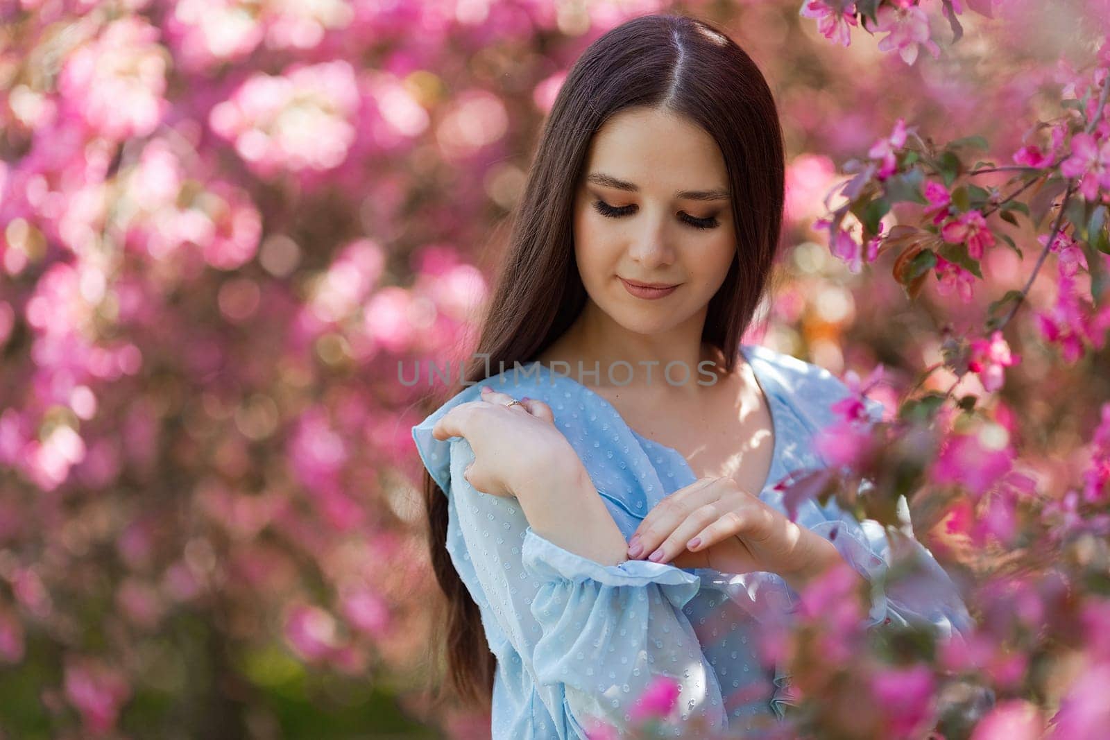 Pretty girl with long hair is standing near a pink blooming apple tree, in the summer in the park. Close up