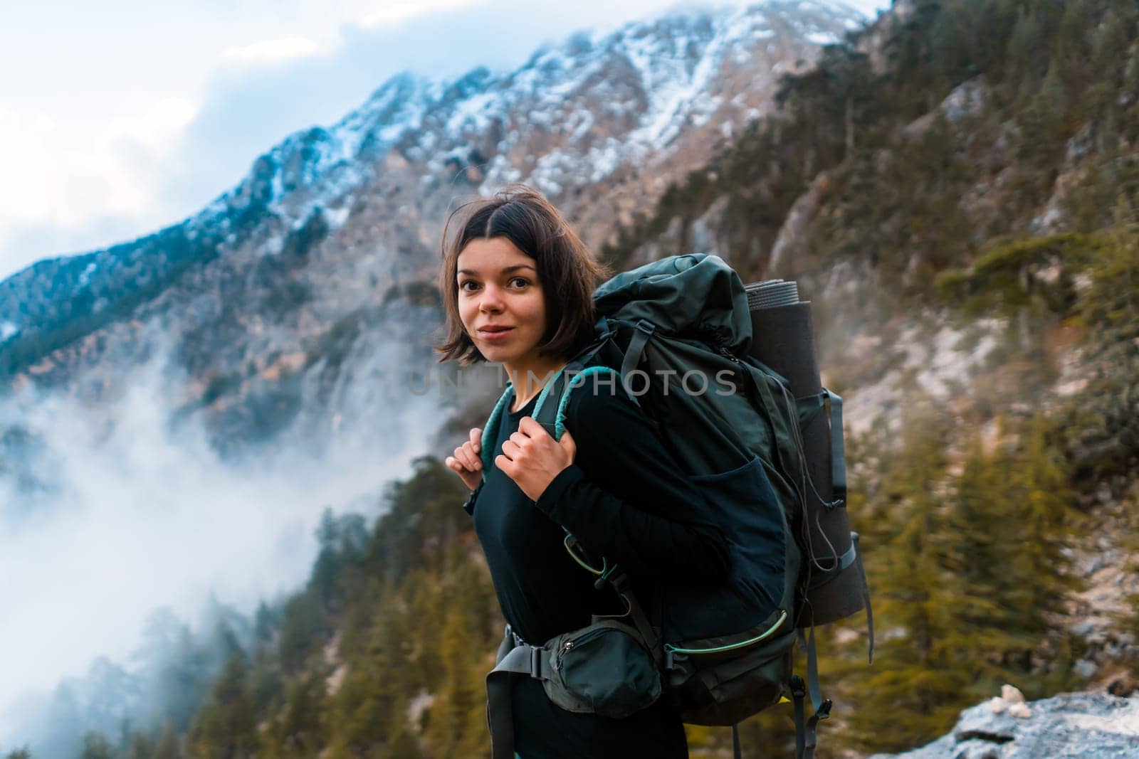 Young smiley girl with backpack and climbing equipment walks on the mountain trail in the national park. The hiker explores wildlife, woodland, beautiful foggy forest and mountains.