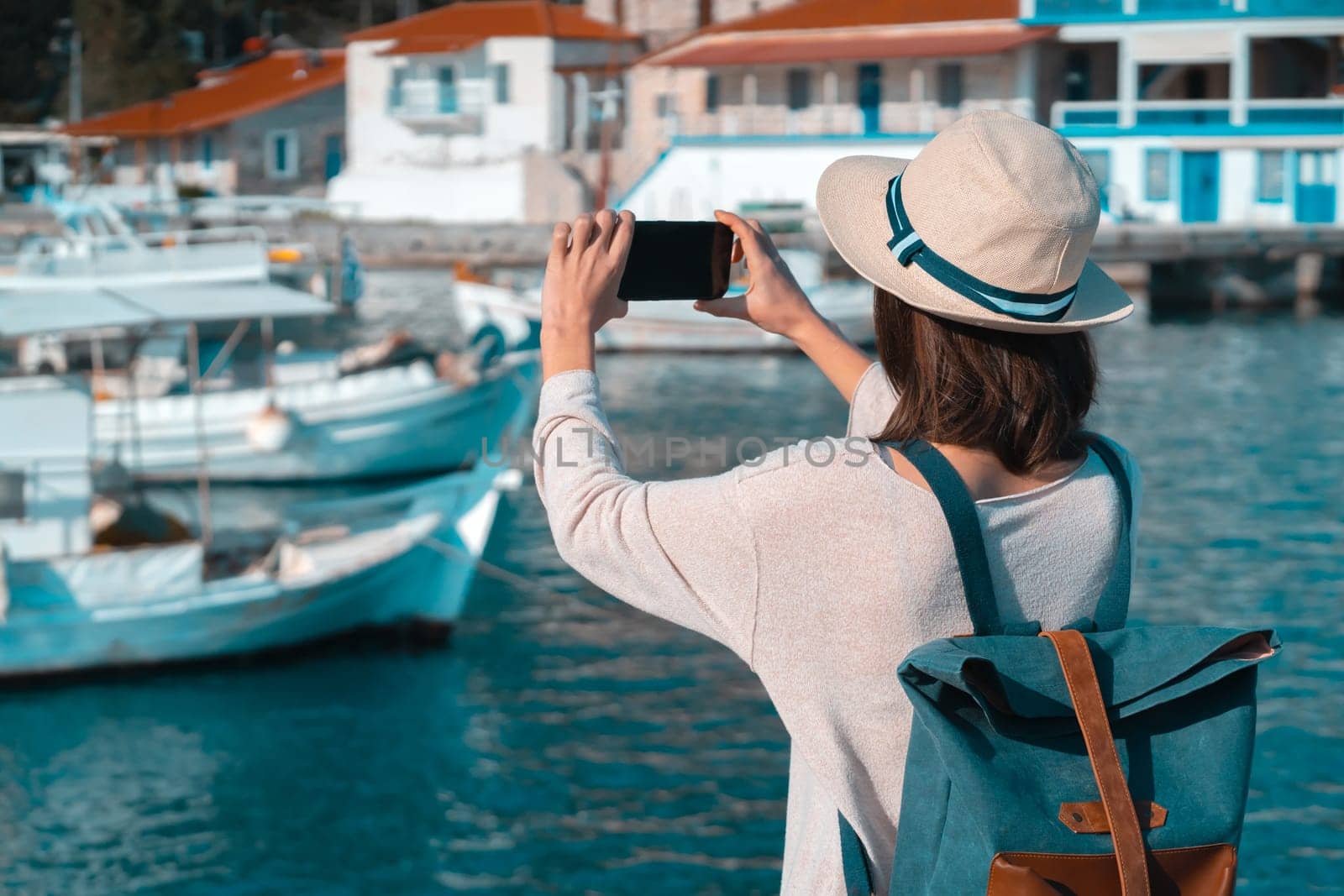 A young girl in a hat and with a backpack makes a photo of a beautiful bay with yachts and boats on the sea while traveling, a tourist walks along the shore next to the ocean and takes pictures.