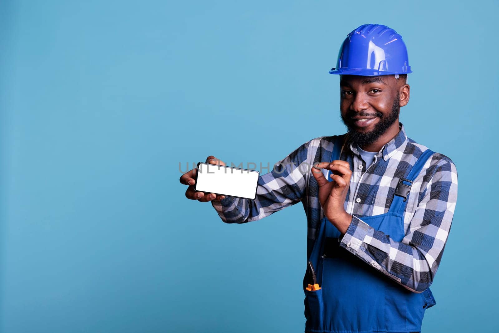 Man in work uniform holding smartphone with blank advertising screen in studio shot. African american construction worker pointing to empty display screen of cell phone on blue background.