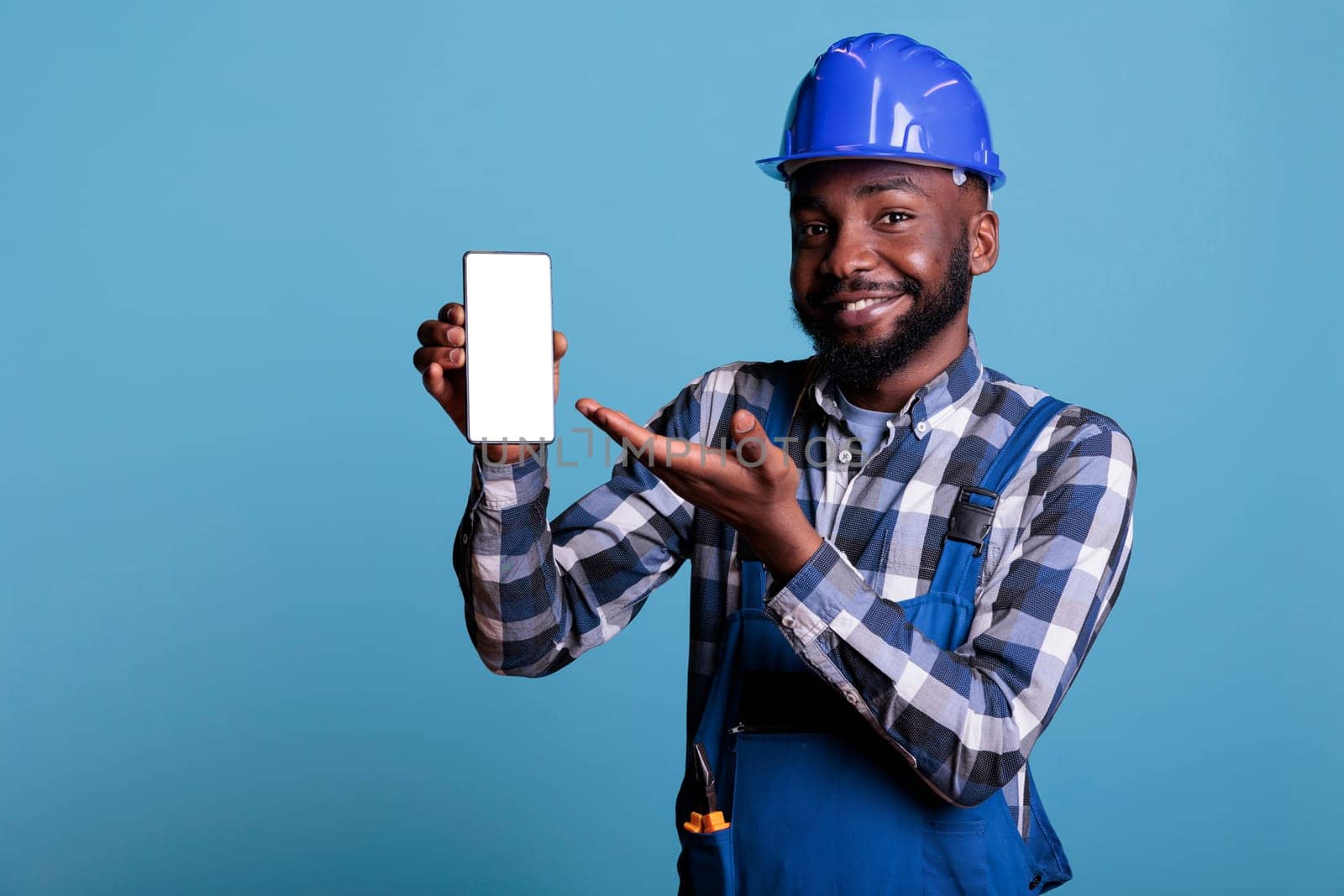 Optimistic african american contractor showing empty mobile phone screen for advertising in studio shot. Construction worker wearing uniform and hard hat against blue background, smiling at camera.
