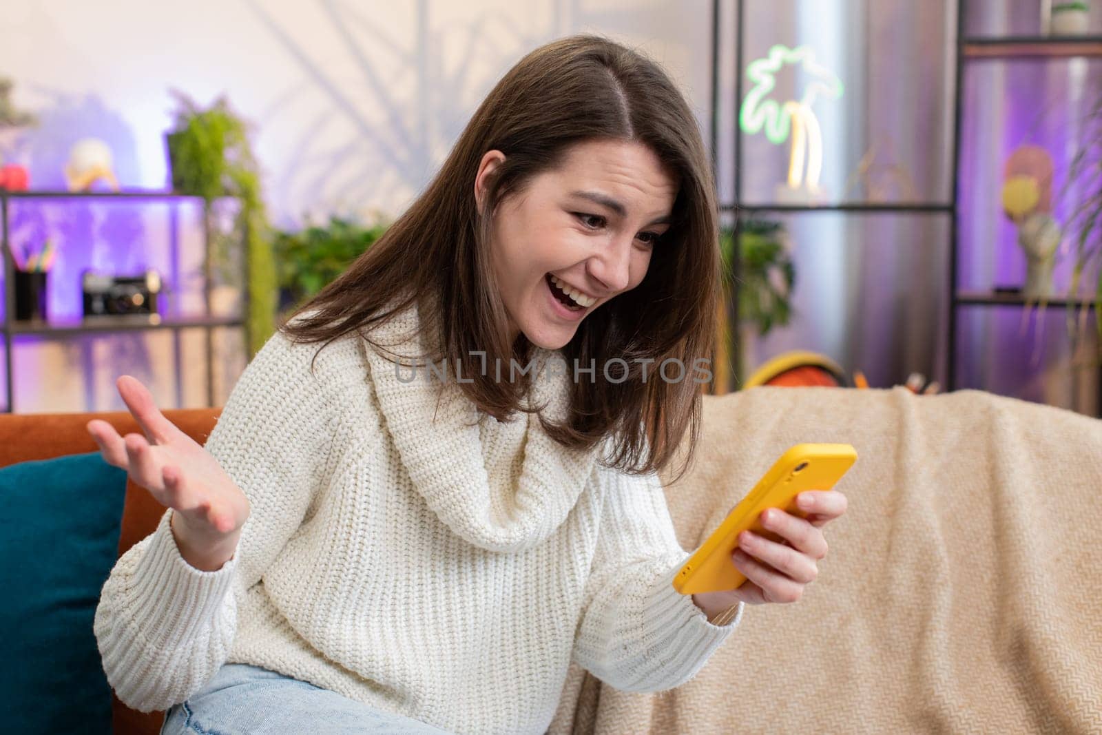 Happy young woman use mobile smartphone typing browsing say Wow yes found out great big win good news celebrate lottery jackpot doing winner gesture. Portrait of girl at home living room sits on sofa