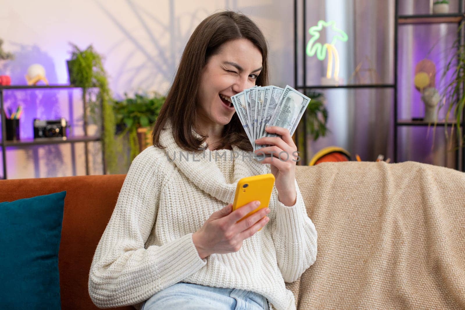 Planning family budget. Smiling young woman counting money cash, use smartphone calculate domestic bills at home room. Joyful girl satisfied of income and saves money for planned vacation, gifts