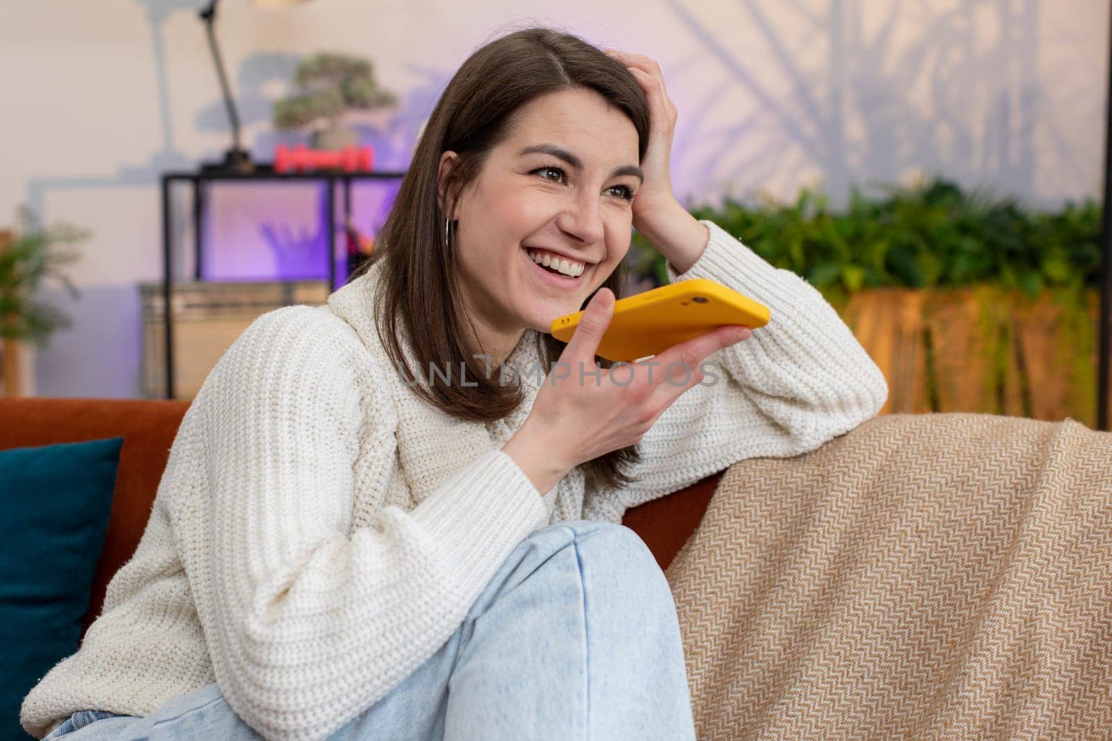 Portrait of happy excited young woman enjoying mobile smartphone loudspeaker talking good news, laughing at home room apartment. Girl making phone call conversation with friends sitting on couch