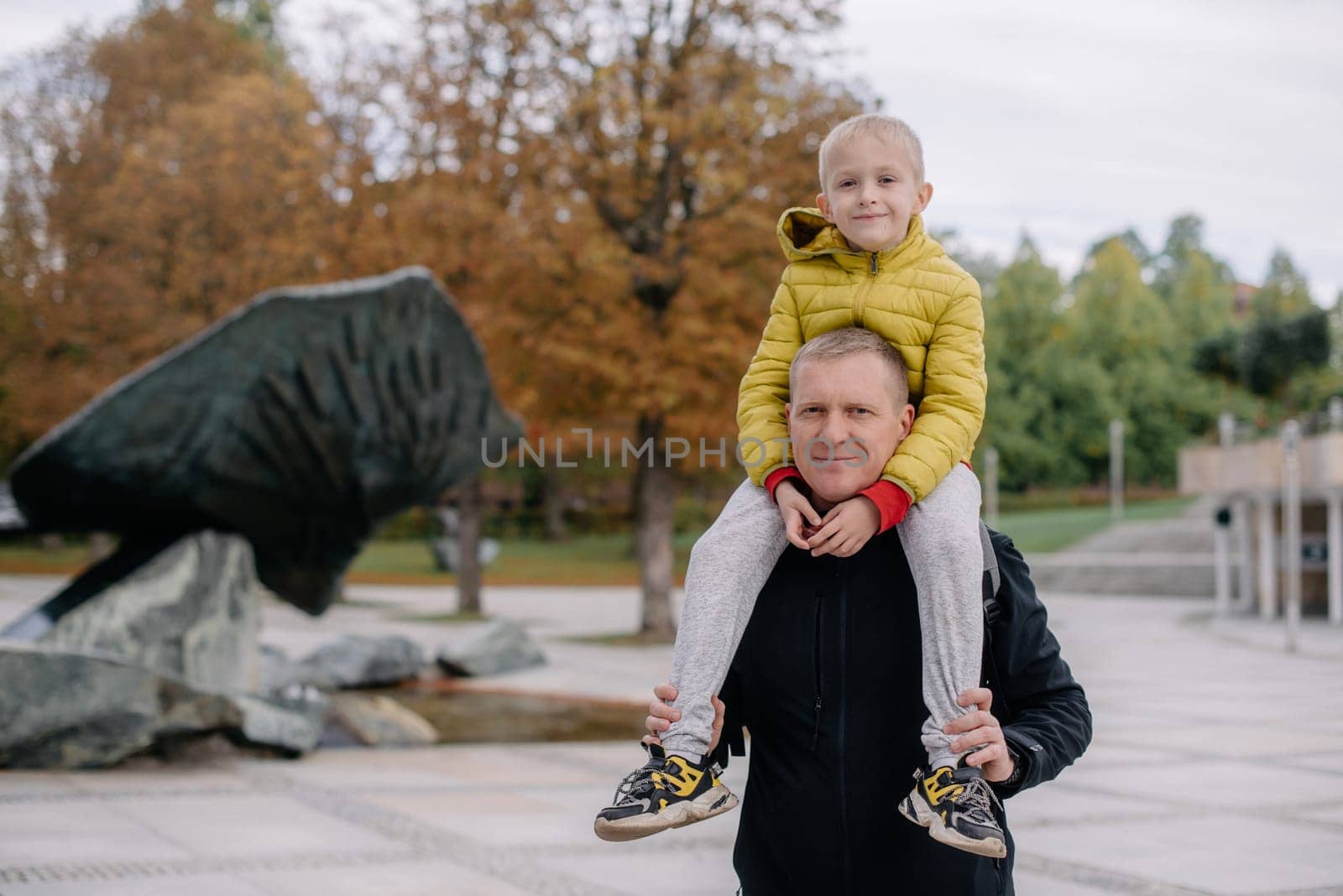 Dad holds on his son's shoulders. Beautiful family is spending time together outside. Dad and his little son are having fun on a roof terrace with view on a city. Sitting on father's shoulders and smiling. by Andrii_Ko