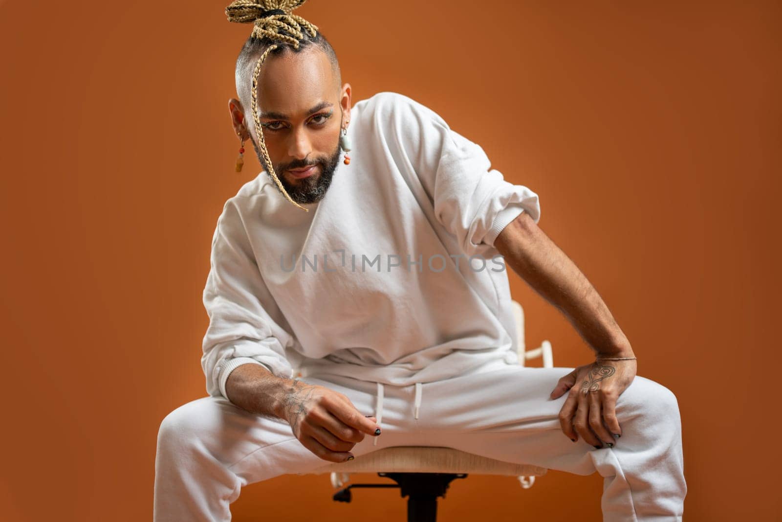 Trendy black latin gay man in white clothes sitting chair look camera isolated on orange background studio portrait People lifestyle fashion lgbtq concept