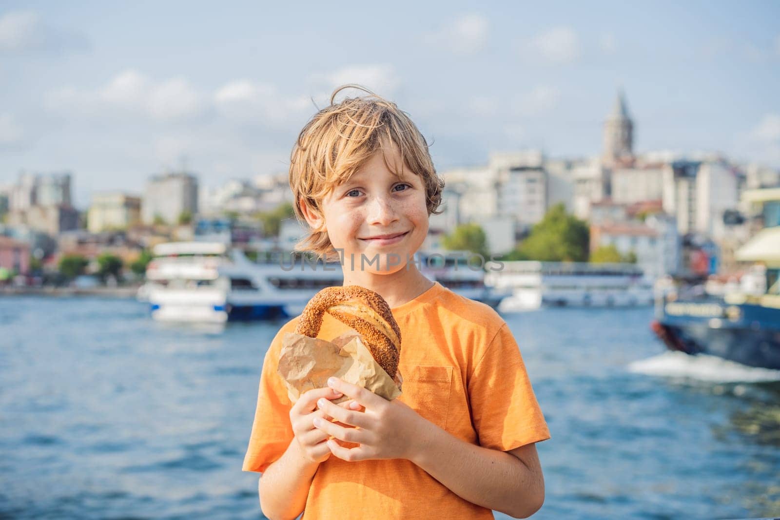 Boy in Istanbul having breakfast with Simit and a glass of Turkish tea. Glass of Turkish tea and bagel Simit against golden horn bay and the Galata Tower in Istanbul, Turkey. Turkiye. Traveling with kids concept.