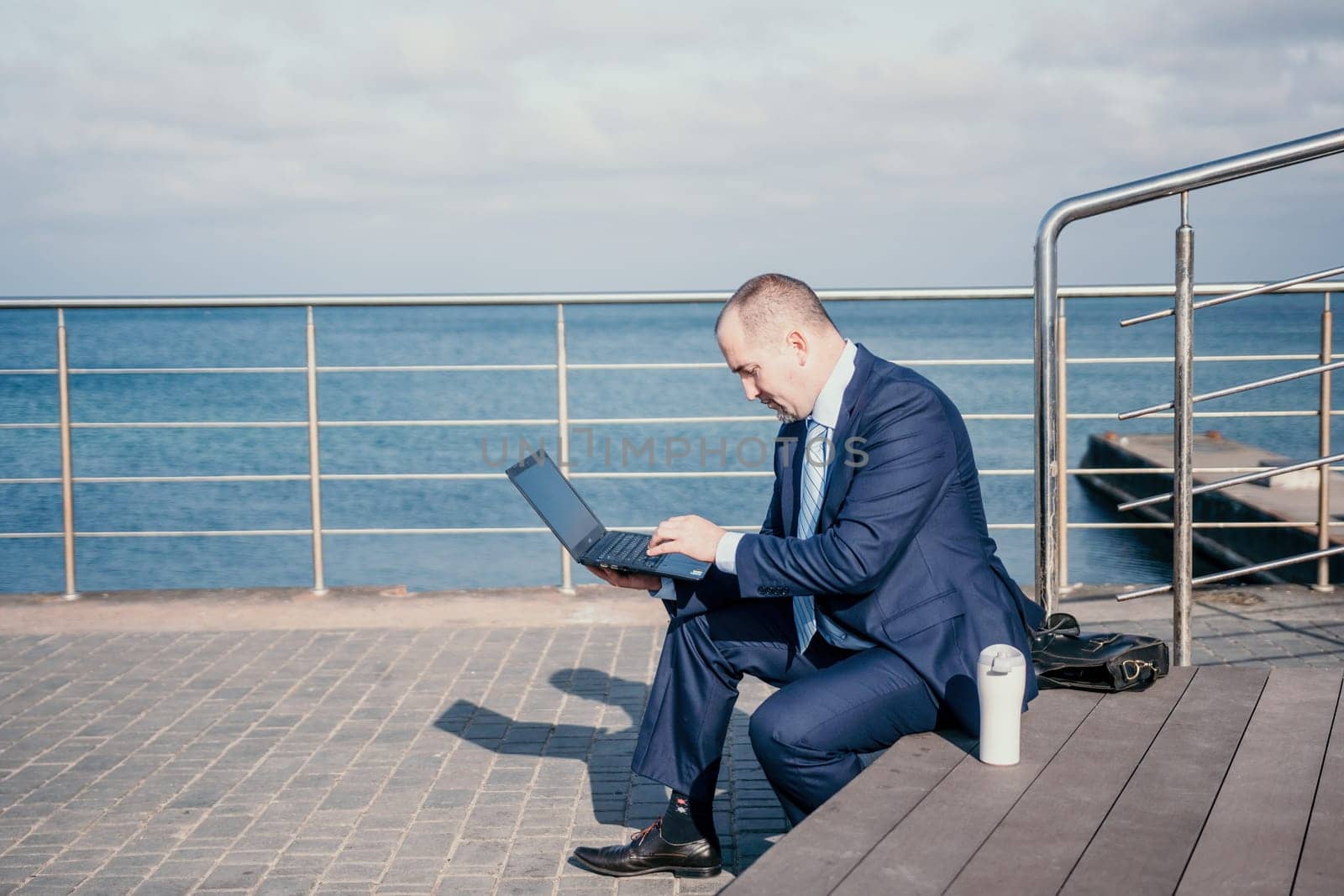 Confident middle age businessman working remotely online, typing on a laptop keyboard while sitting on a beach at sunset. Working remotely on vacation, running an online business from a distance. by panophotograph