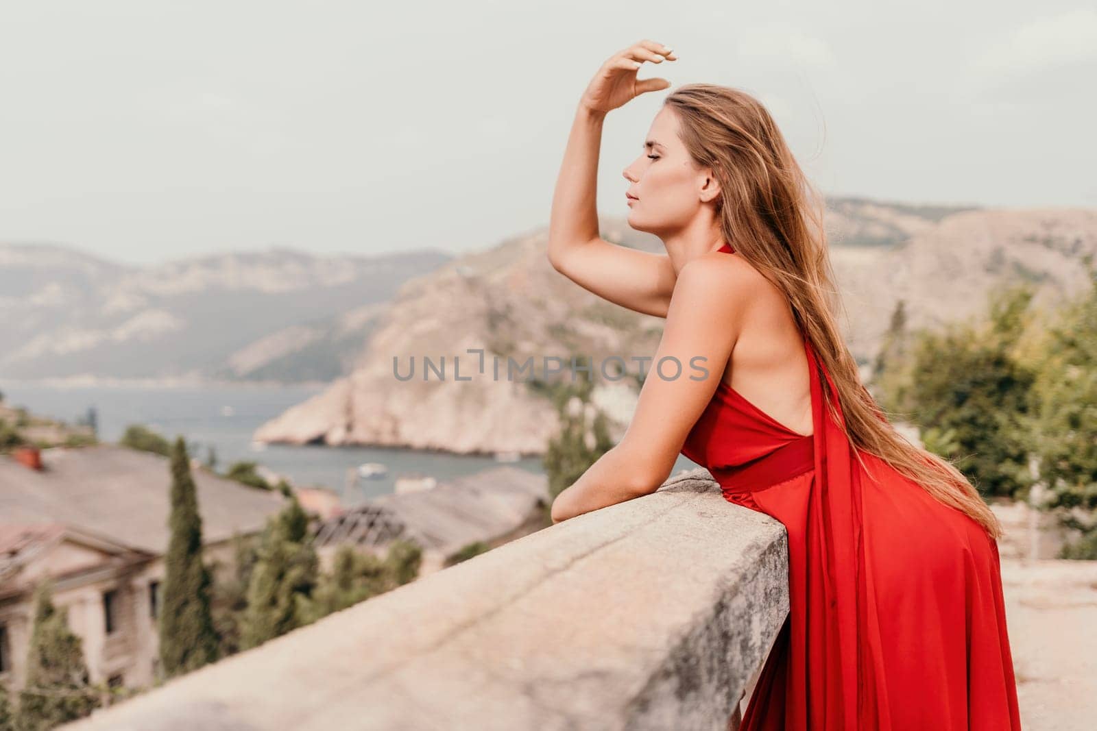 Side view a Young beautiful sensual woman in a red long dress posing on a volcanic rock high above the sea during sunset. Girl on the nature on overcast sky background. Fashion photo