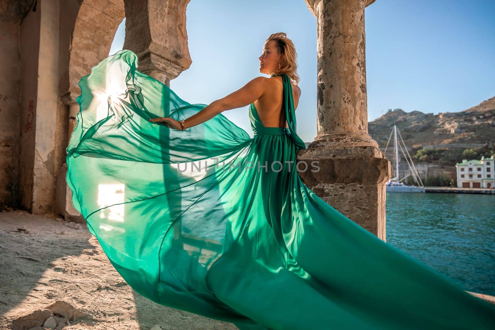 Rear view of a happy blonde woman in a long mint dress posing against the backdrop of the sea in an old building with columns. Girl in nature against the blue sky
