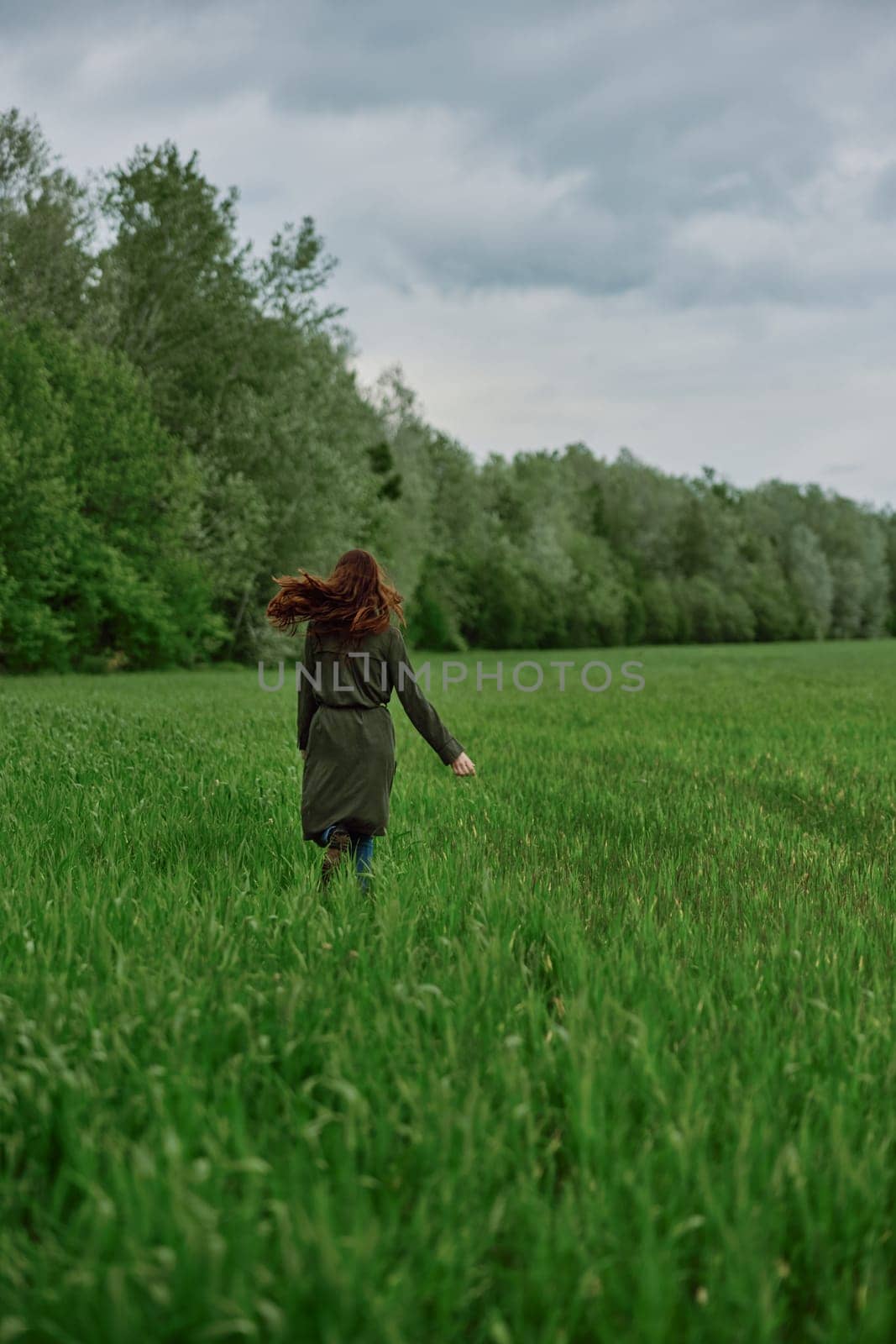 a woman in a long raincoat runs across a field in tall green grass in cloudy weather in spring by Vichizh