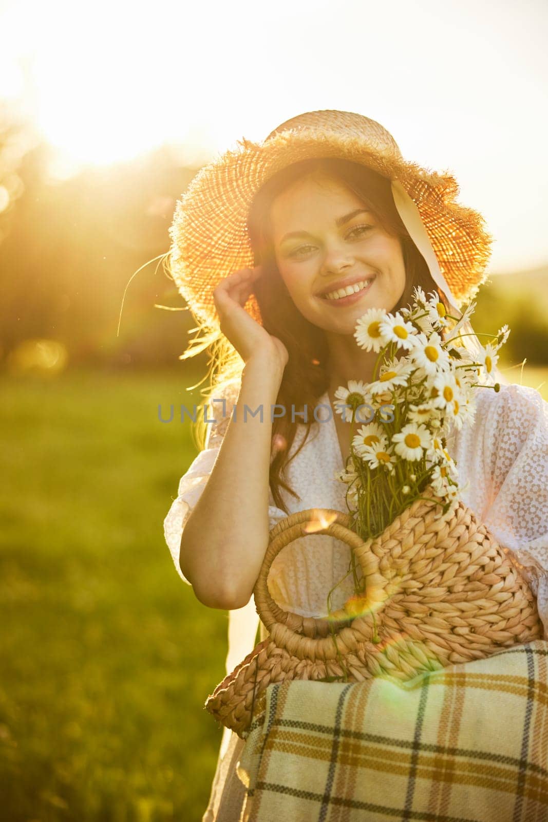 portrait of a happily smiling woman in a field lit from the back by the rays of the setting sun with a basket of flowers and a blanket in her hands. High quality photo