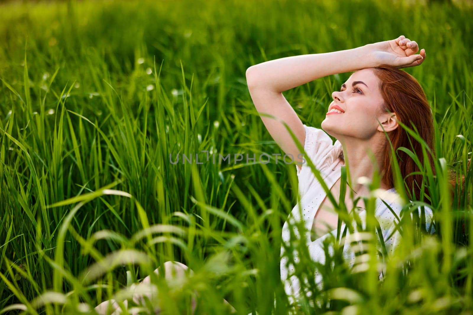 cute woman with red hair sits in tall green grass with her hand on her head. High quality photo