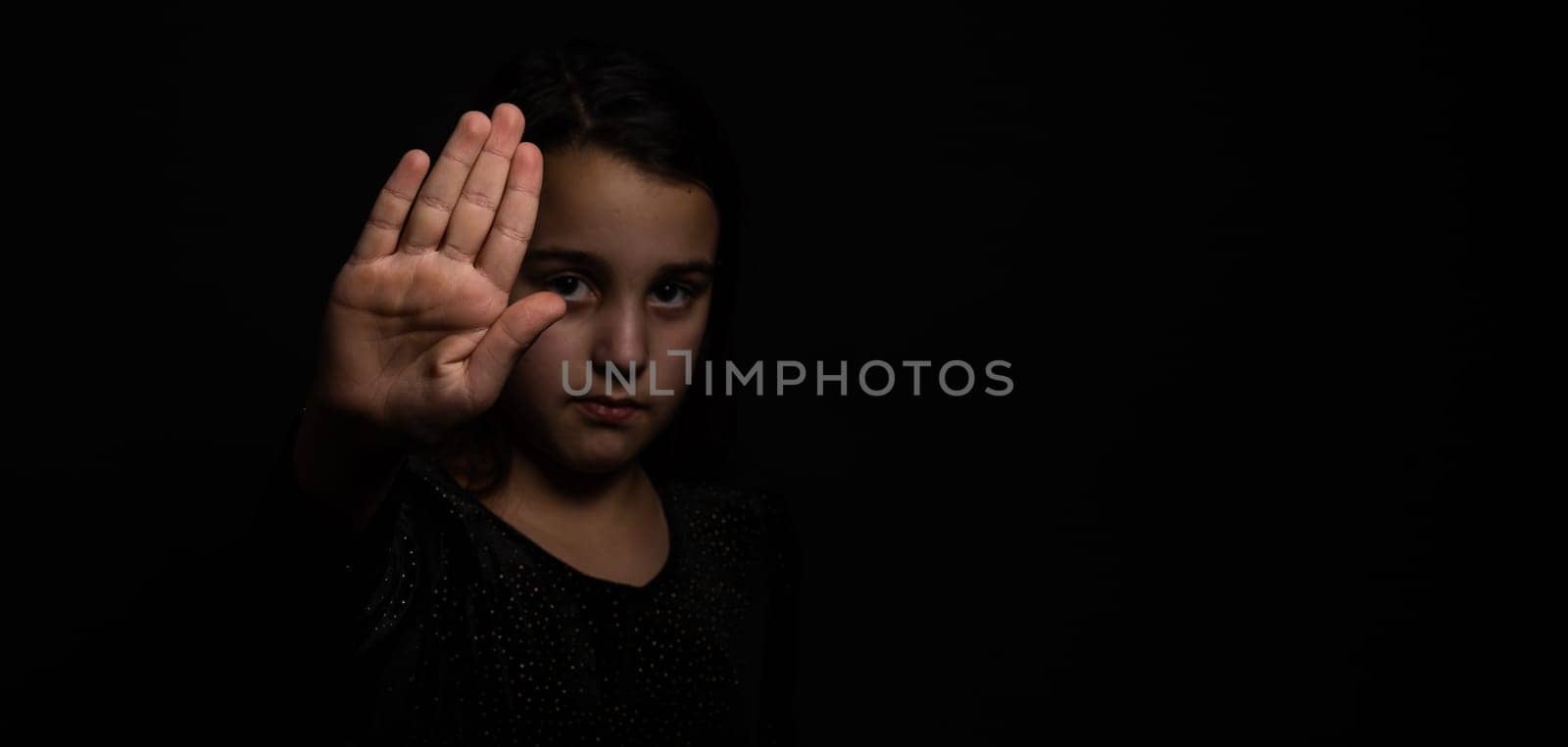 little girl with a raised hand making a stop sign gesture on a black background. Violence, harassment and child abuse prevention concept. by Andelov13