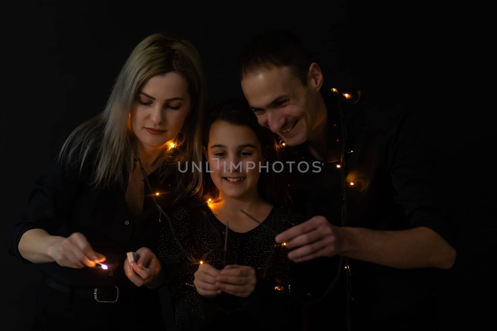 Happy family in black - woman, man and little girl, with a sparkler in hand the New Year's Christmas at home. The girl at the father on hands. New year 2023