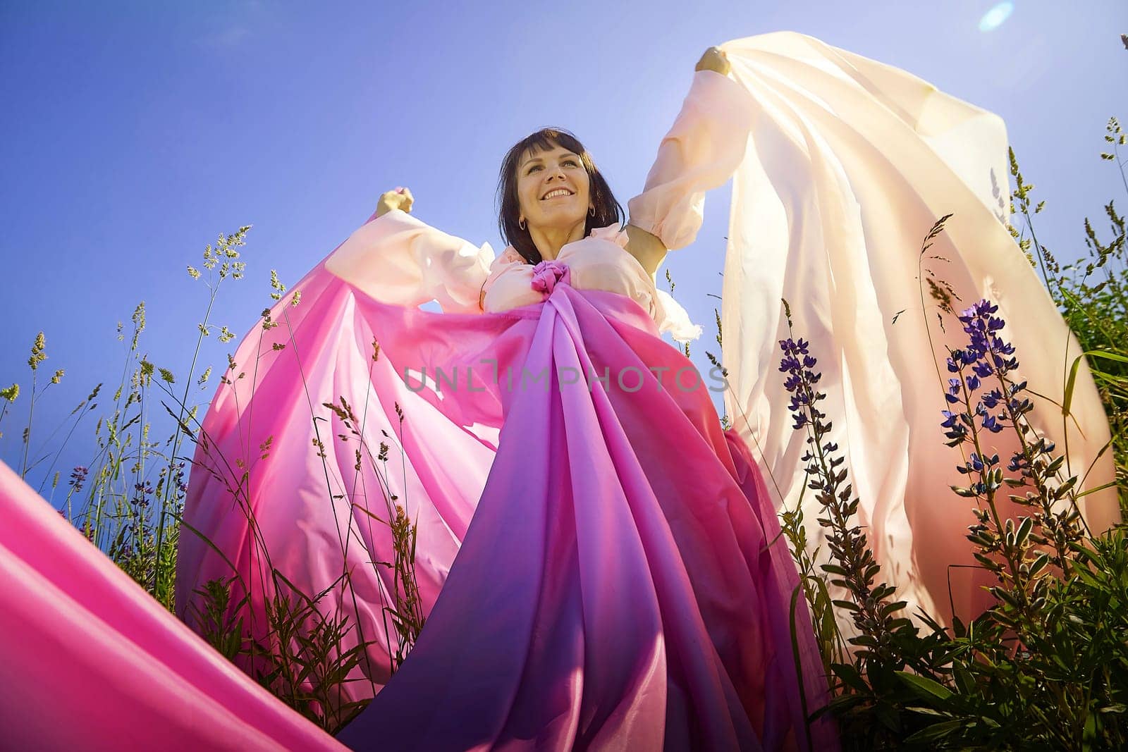 Beautiful girl in a lush pink ball gown in green field during blooming of flowers and blue sky on background. Model posing on nature landscape as princess from fary tale by keleny