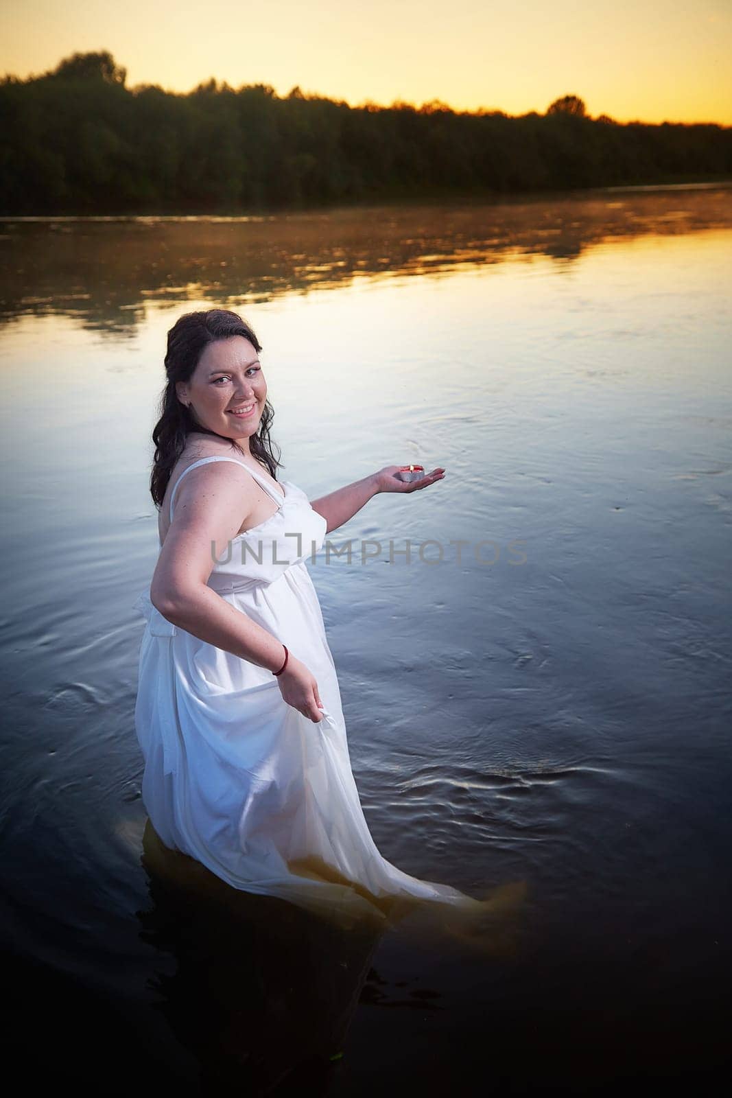 Slavic plump plump chubby girl in long white dress on the feast of Ivan Kupala with flowers and water in a river or lake on summer evening