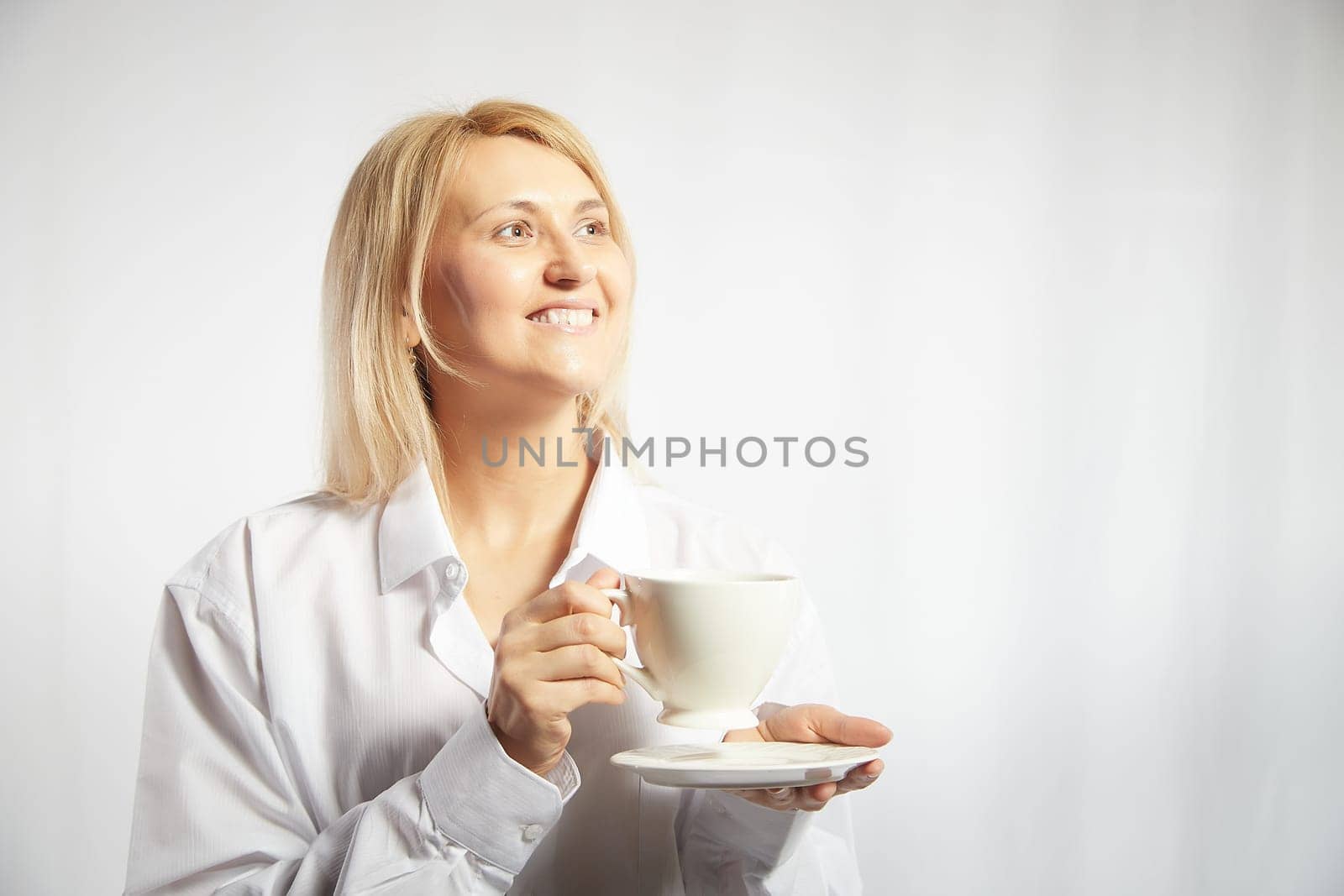 Portrait of pretty blonde smiling woman posing with cup of tea or coffee on white background. Happy girl model in white shirt in studio. The concept of pleasant morning at home or at work. Copy space by keleny