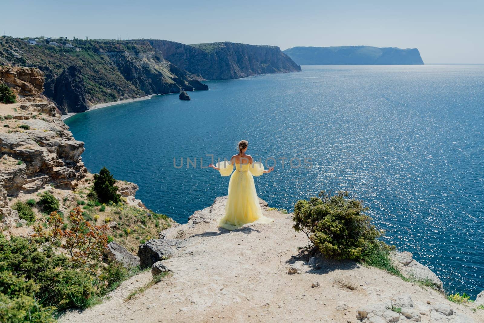 Woman in a yellow dress on the sea. Side view Young beautiful sensual woman in yellow long dress posing on a rock high above the sea at sunset. Girl in nature against the blue sky by Matiunina