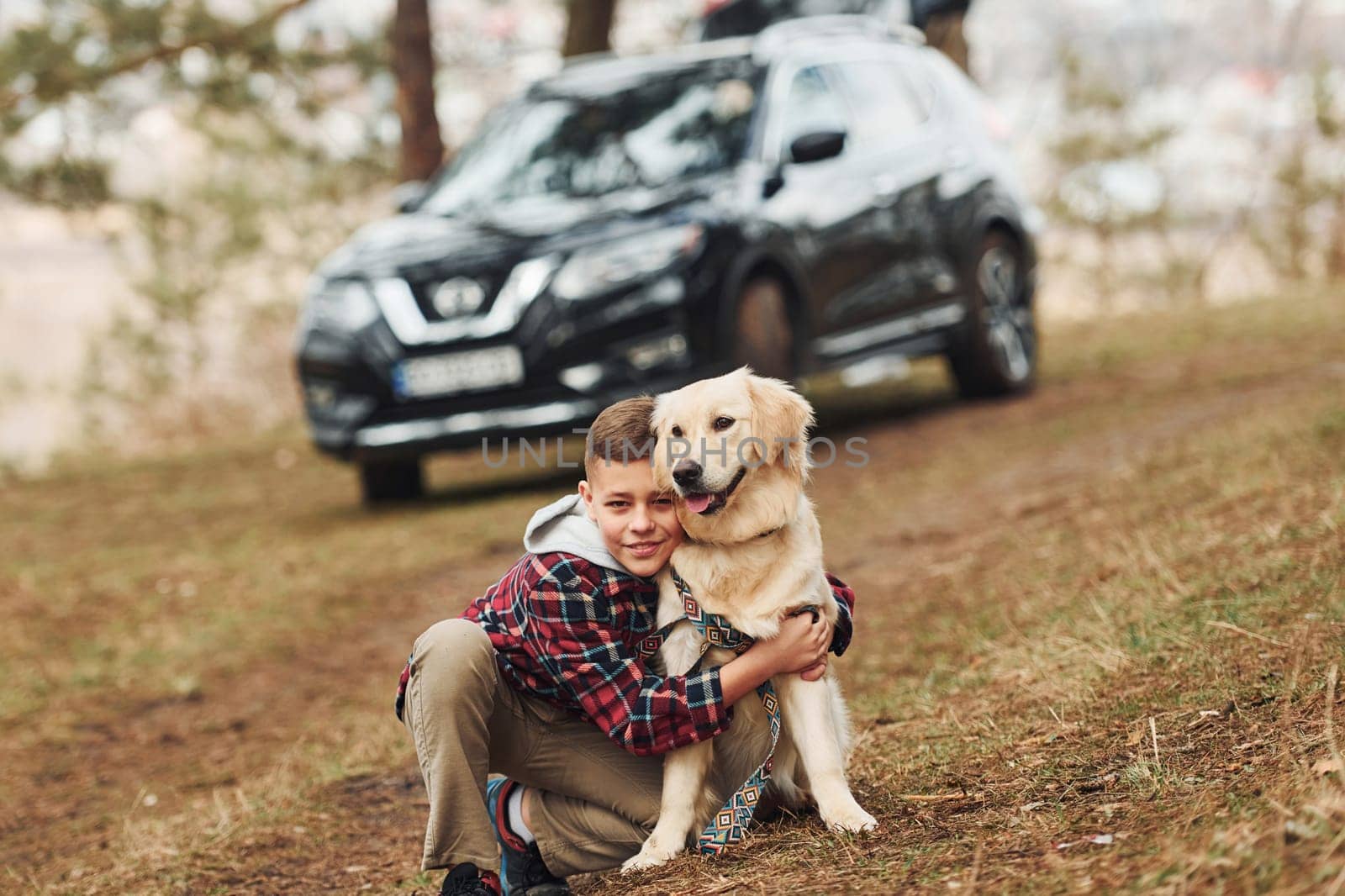 Cheerful boy in casual clothes sitting with her dog in forest against modern black car by Standret