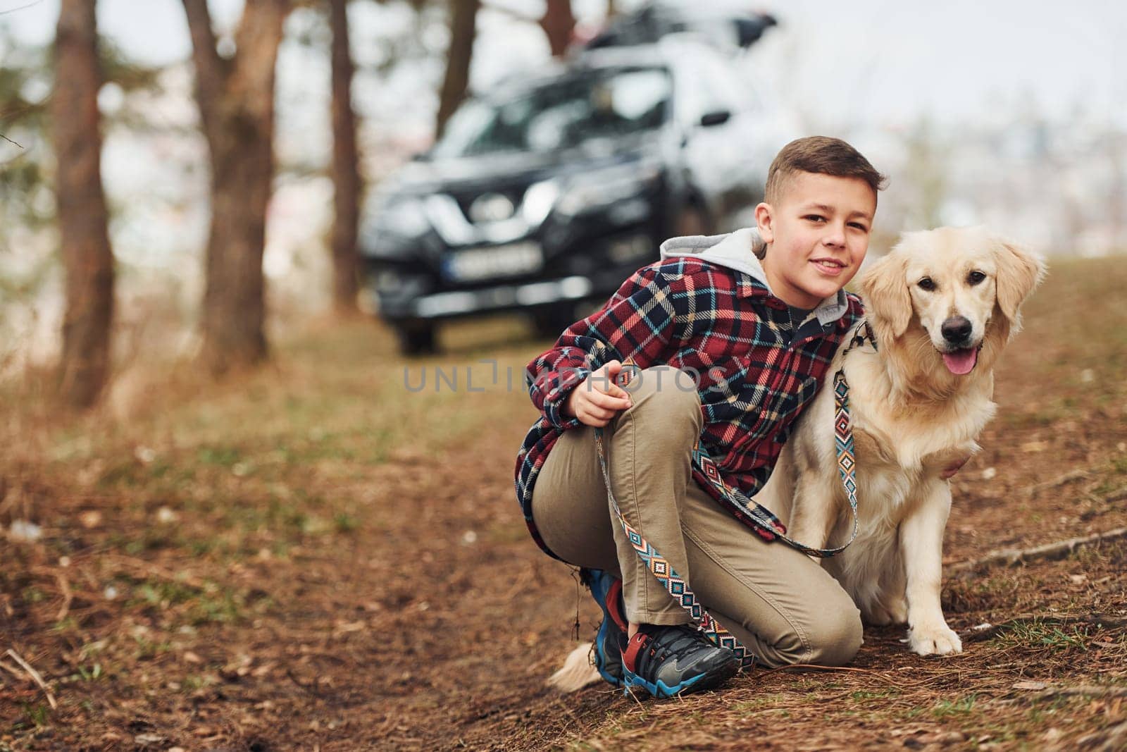 Cheerful boy in casual clothes sitting with her dog in forest against modern black car.
