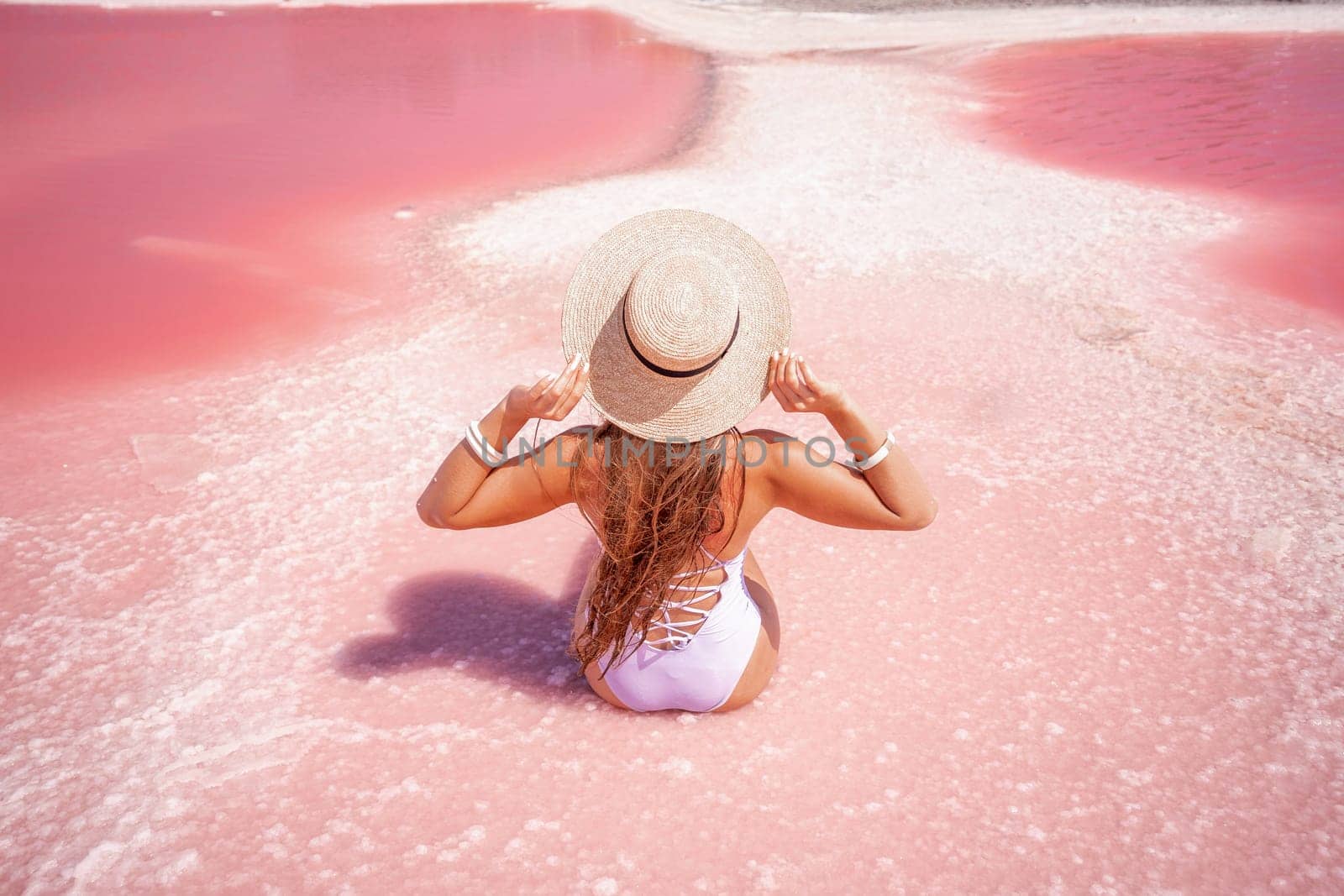 A woman traveler looks at an amazing pink salt lake. He sits with his back in a bathing suit and holds his hat in his hands. Wanderlust travel concept.