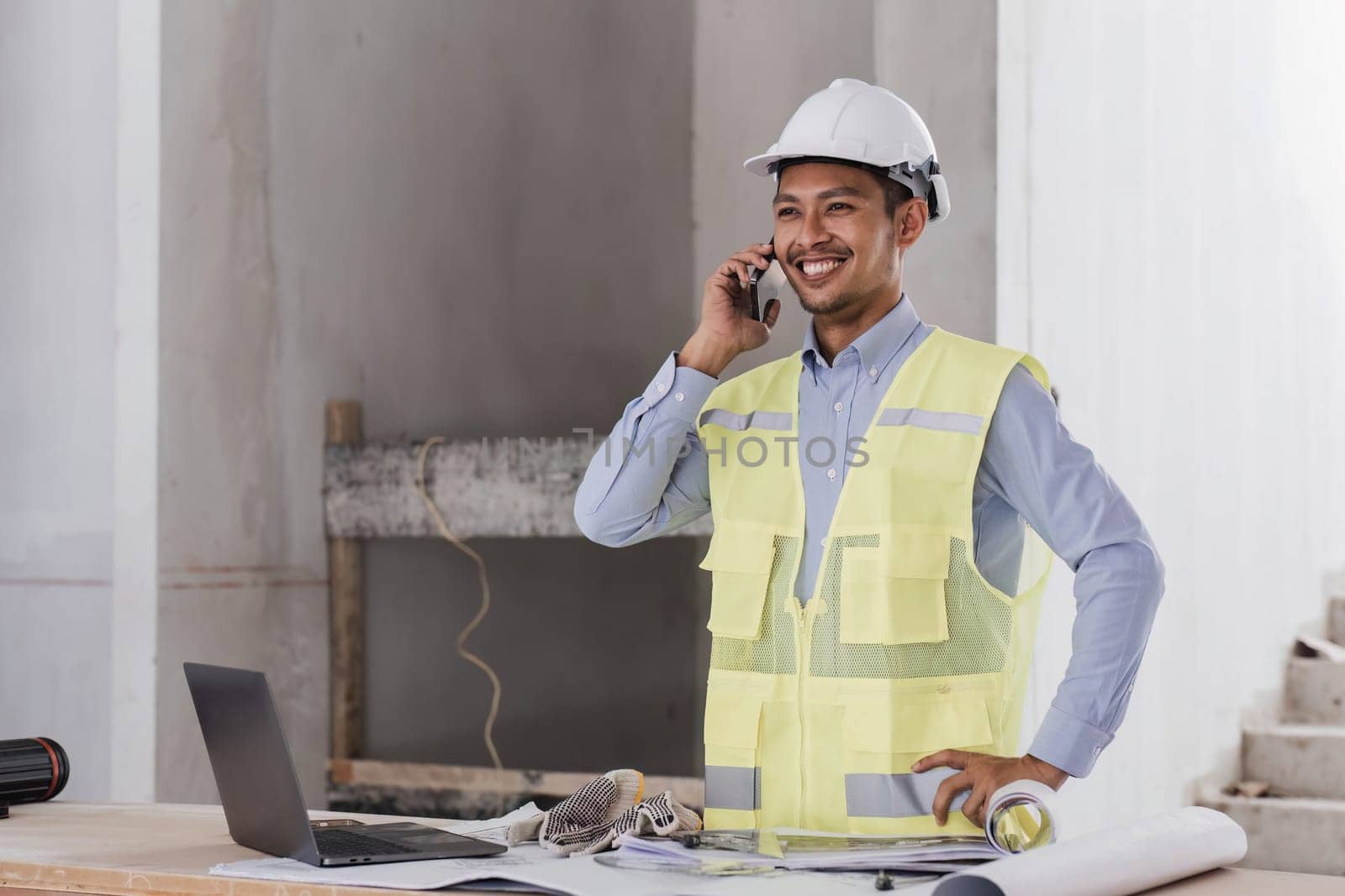 Engineer or Architect with protective safety helmet checking using smartphone holding architectural drawing at construction site. Engineering, Architecture and building project concepts..