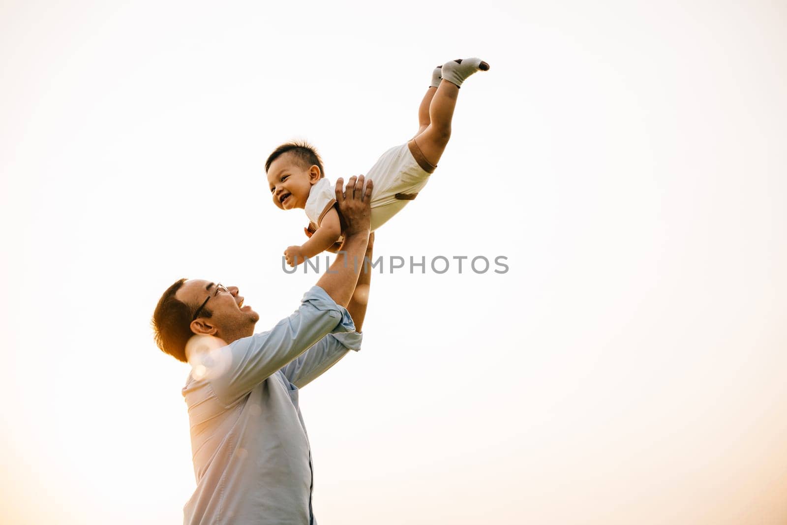 In a beautiful moment of family togetherness, a father holds his baby daughter up high, throwing her up into the sunny sky at sunset. The cheerful child smiles with freedom and playful fun