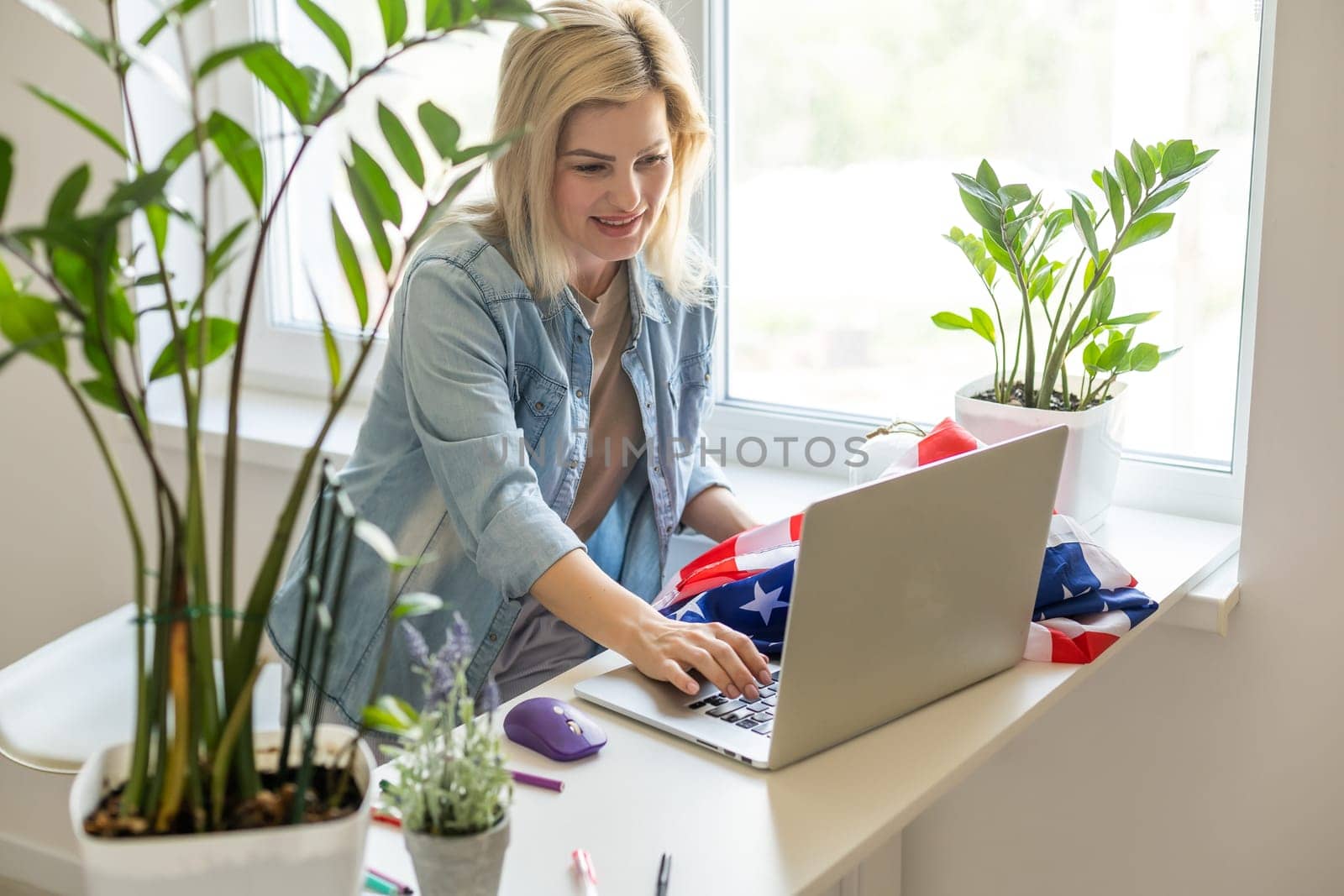 Happy woman employee sitting wrapped in USA flag, shouting for joy in office workplace, celebrating labor day or US Independence day. Indoor studio studio shot isolated on yellow background.