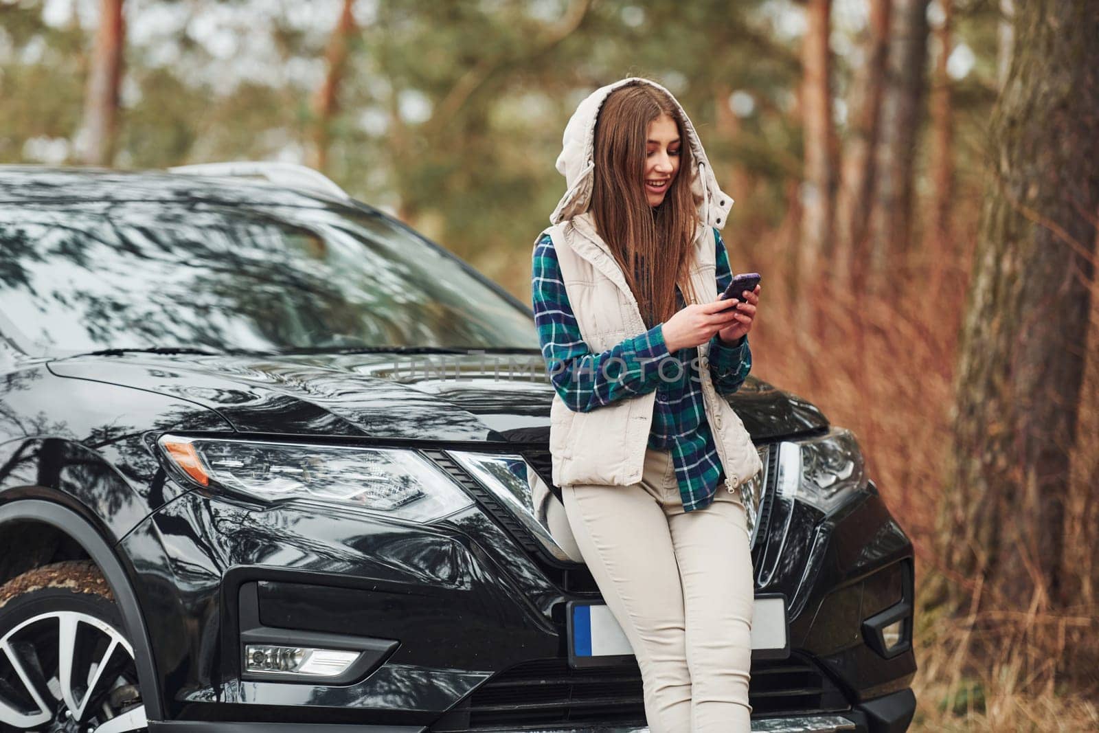 Young cheerful girl standing near modern black car outdoors in the forest.