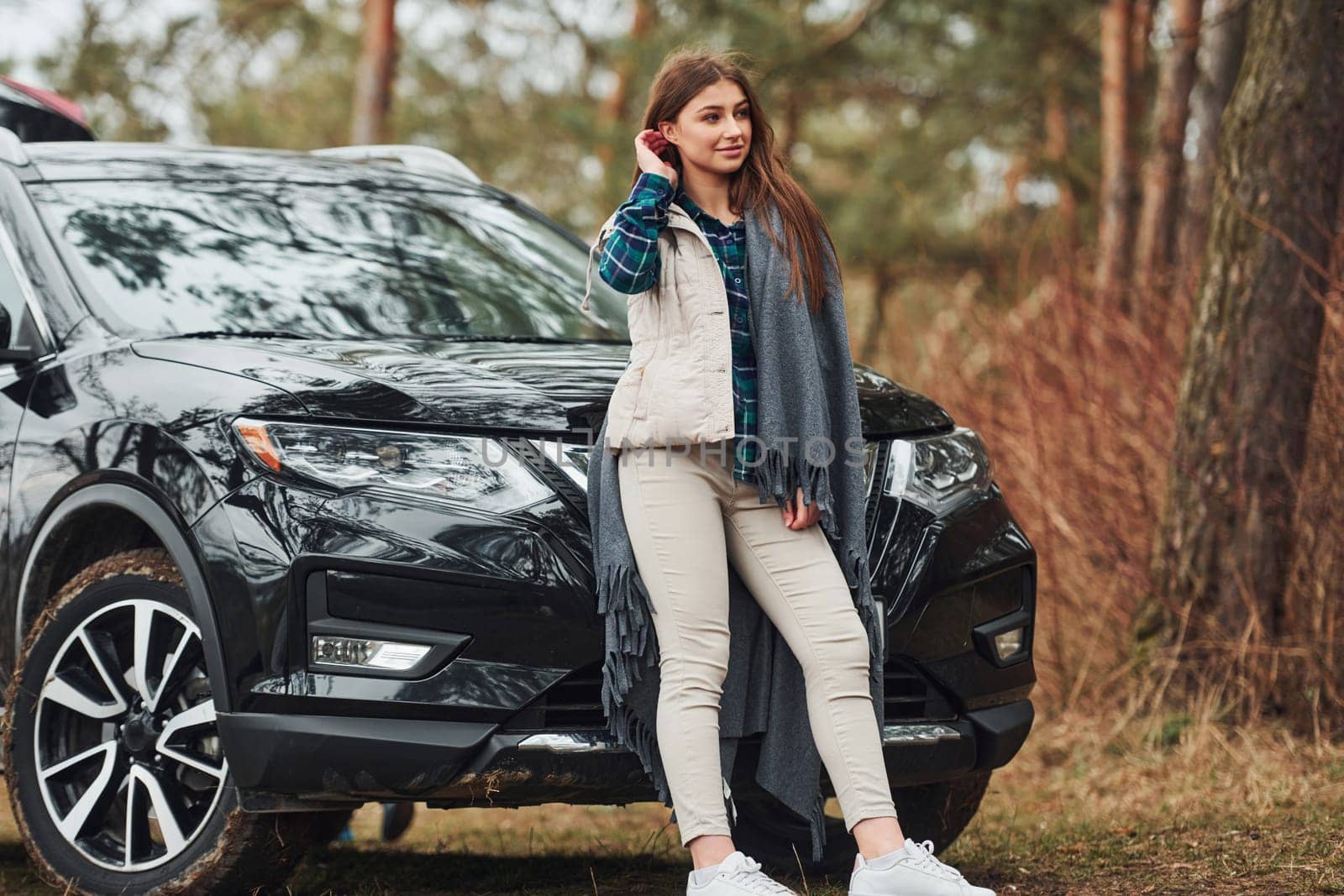 Young cheerful girl standing near modern black car outdoors in the forest by Standret