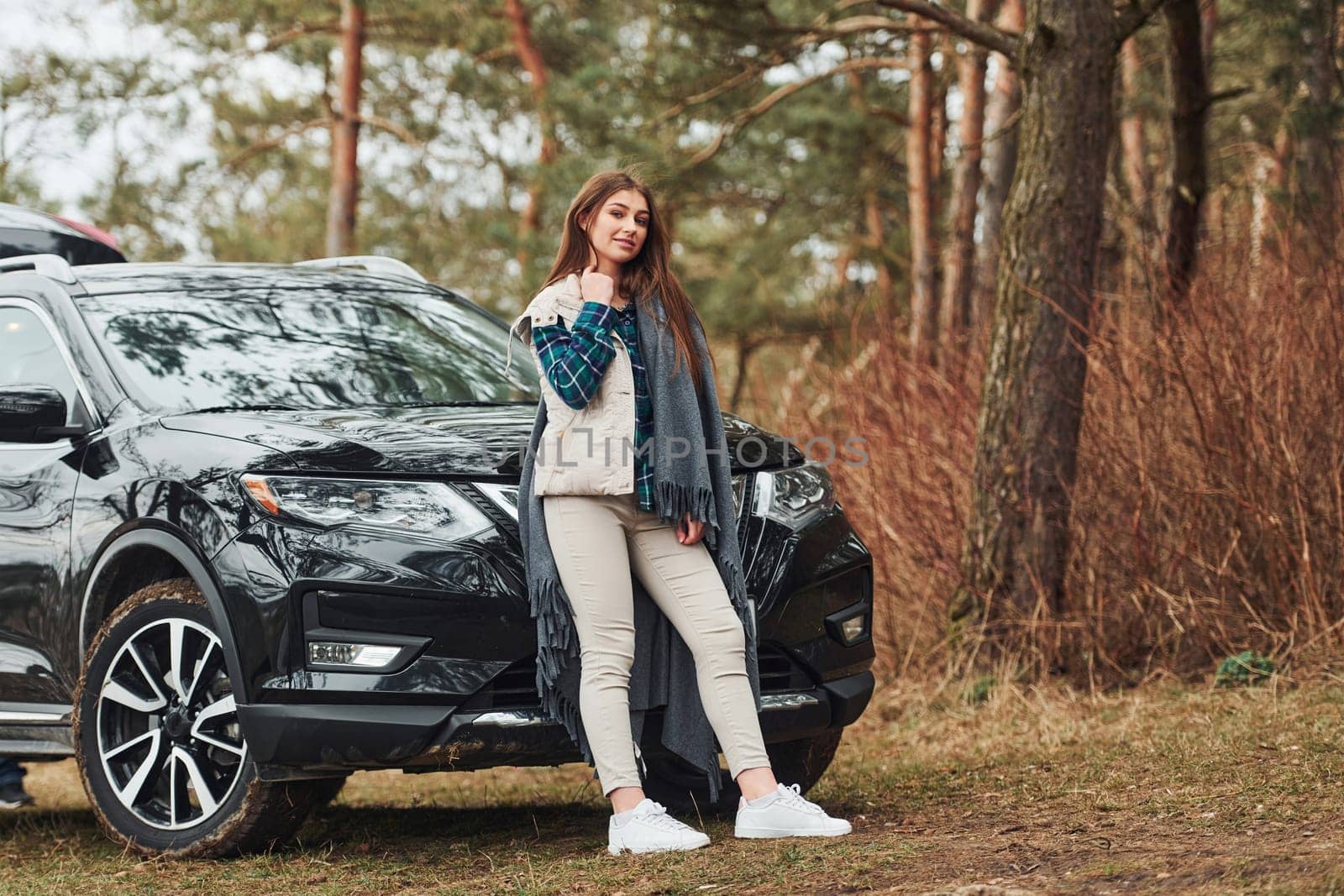 Young cheerful girl standing near modern black car outdoors in the forest.