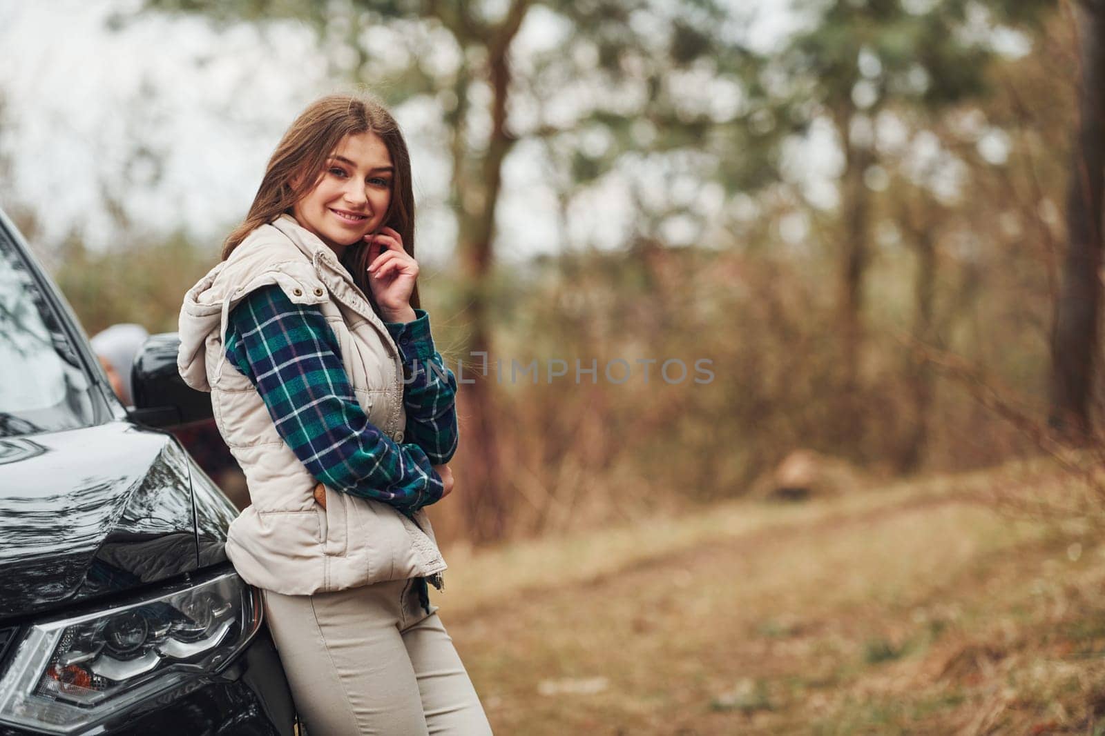 Young cheerful girl standing near modern black car outdoors in the forest.