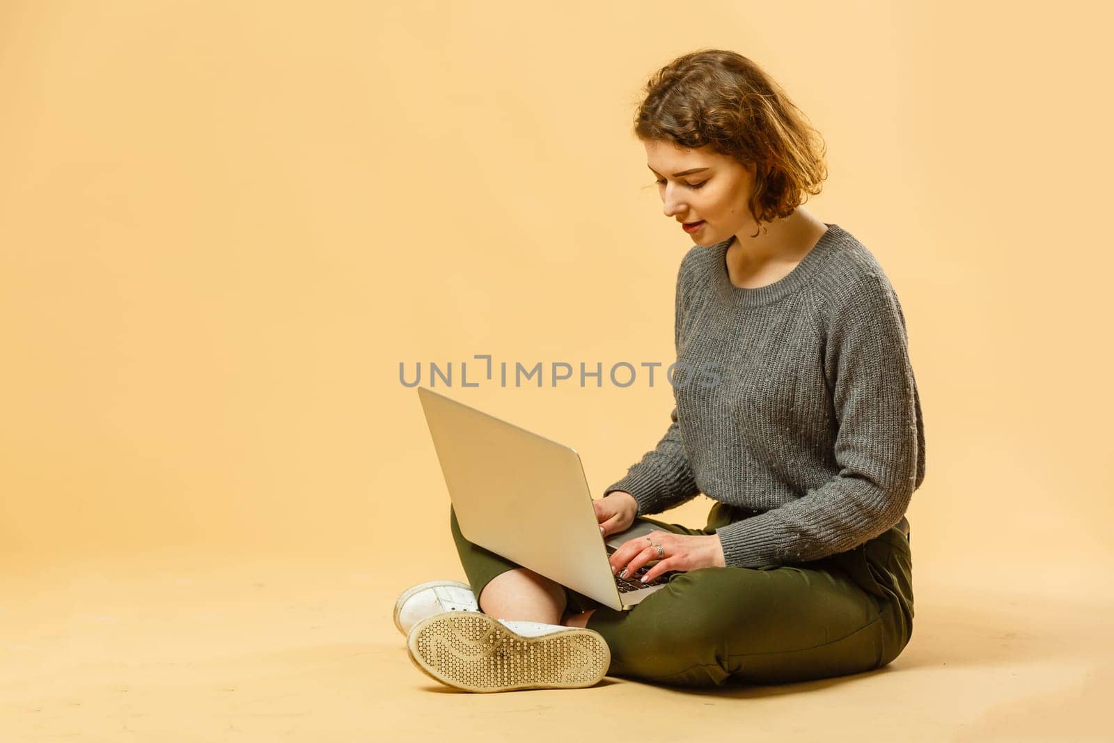 Smiling cheerful funny beautiful attractive young brunette woman 20s wearing basic casual working on laptop pc computer isolated on bright beige colour background studio portrait by Andelov13