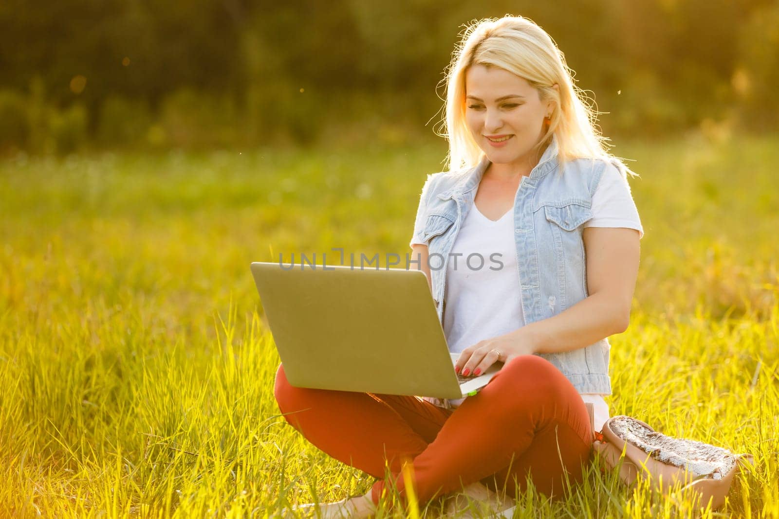 Young woman with laptop sits on the grass in the park on a sunny day. Laughing blonde in a white tank top and shorts. Online training, remote work and communication in social networks.