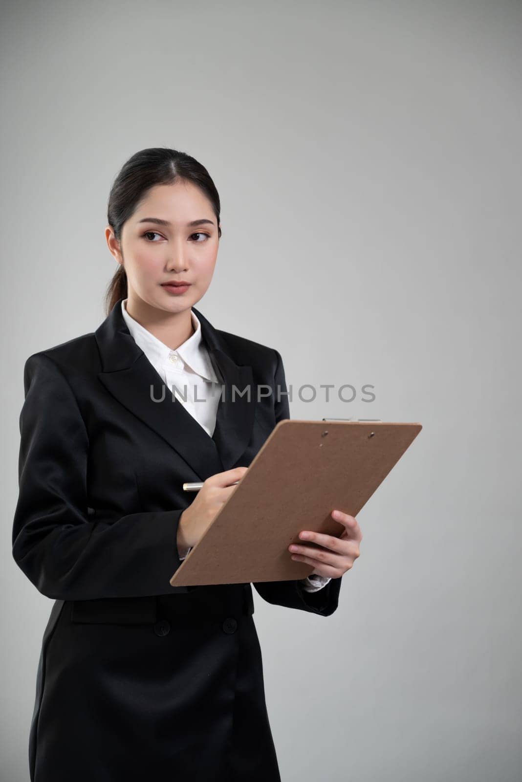 Confident young businesswoman stands on isolated background, holding clipboard and posing in formal black suit. Office lady or manager with smart and professional appearance. Enthusiastic