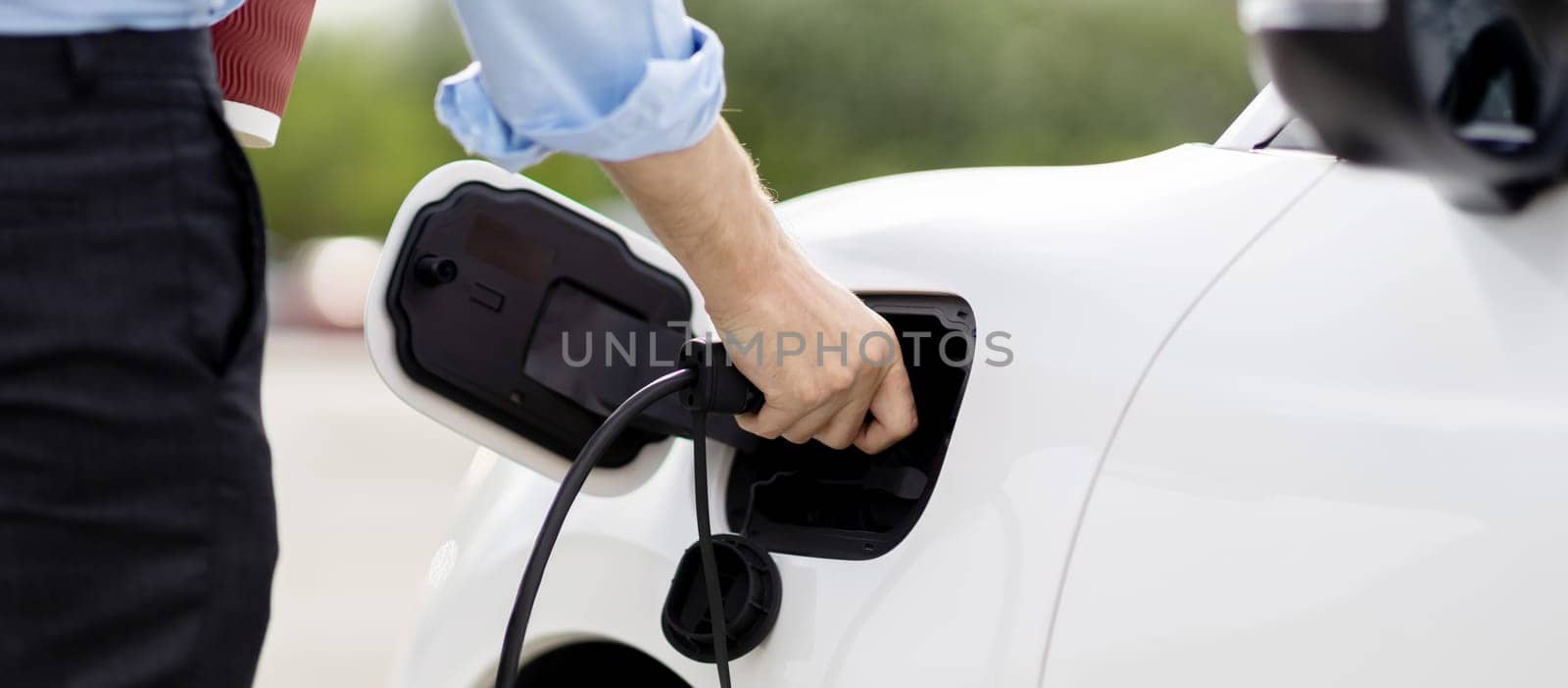 Closeup progressive man holding EV charger plug from public charging station for electric vehicle with background of residential building as concept eco-friendly sustainability energy car concept.