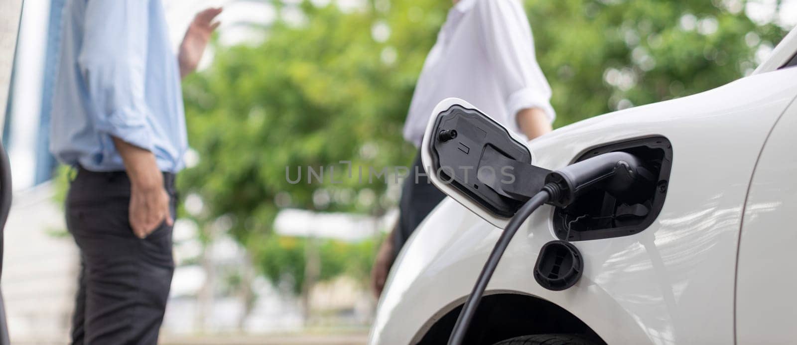 Focus parking-electric car connected to public charging station with blur progressive businesspeople holding coffee, residential building apartment and condo background for eco-friendly concept.