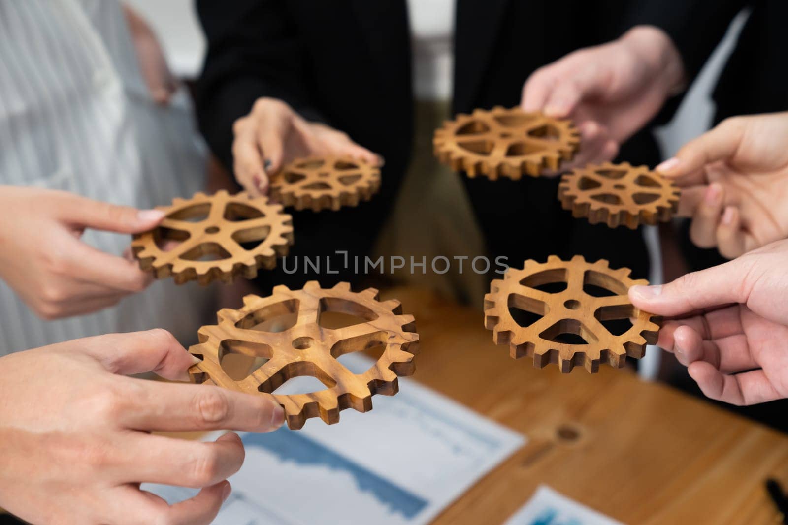 Closeup top view businesspeople hand holding gear and join together over meeting table with financial report papers. Cohesive group of office workers holding cog wheel as synergy harmony concept.