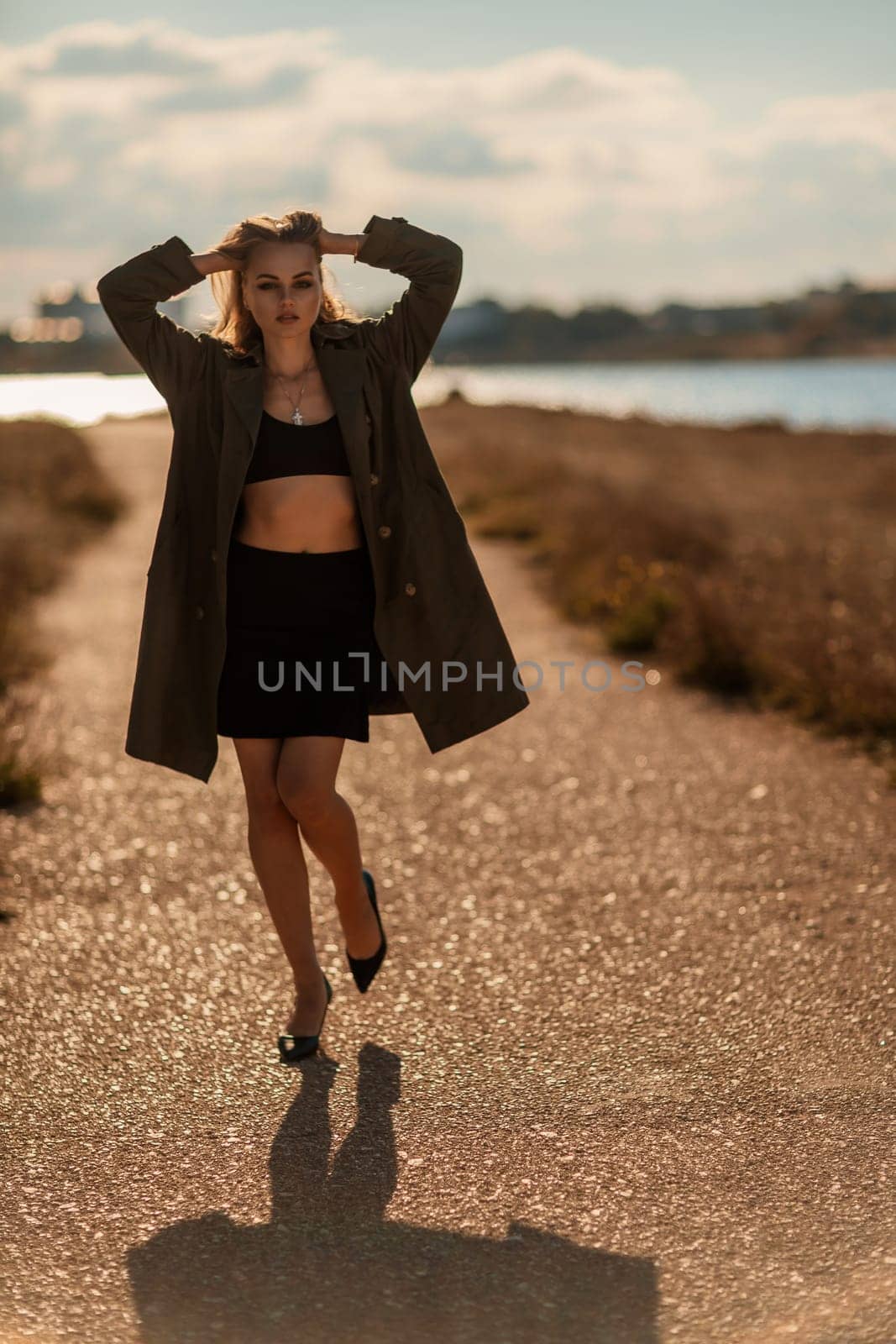 Portrait blonde sea cape. A calm young blonde in an unbuttoned khaki raincoat walks along the seashore, under a raincoat a black skirt and top.